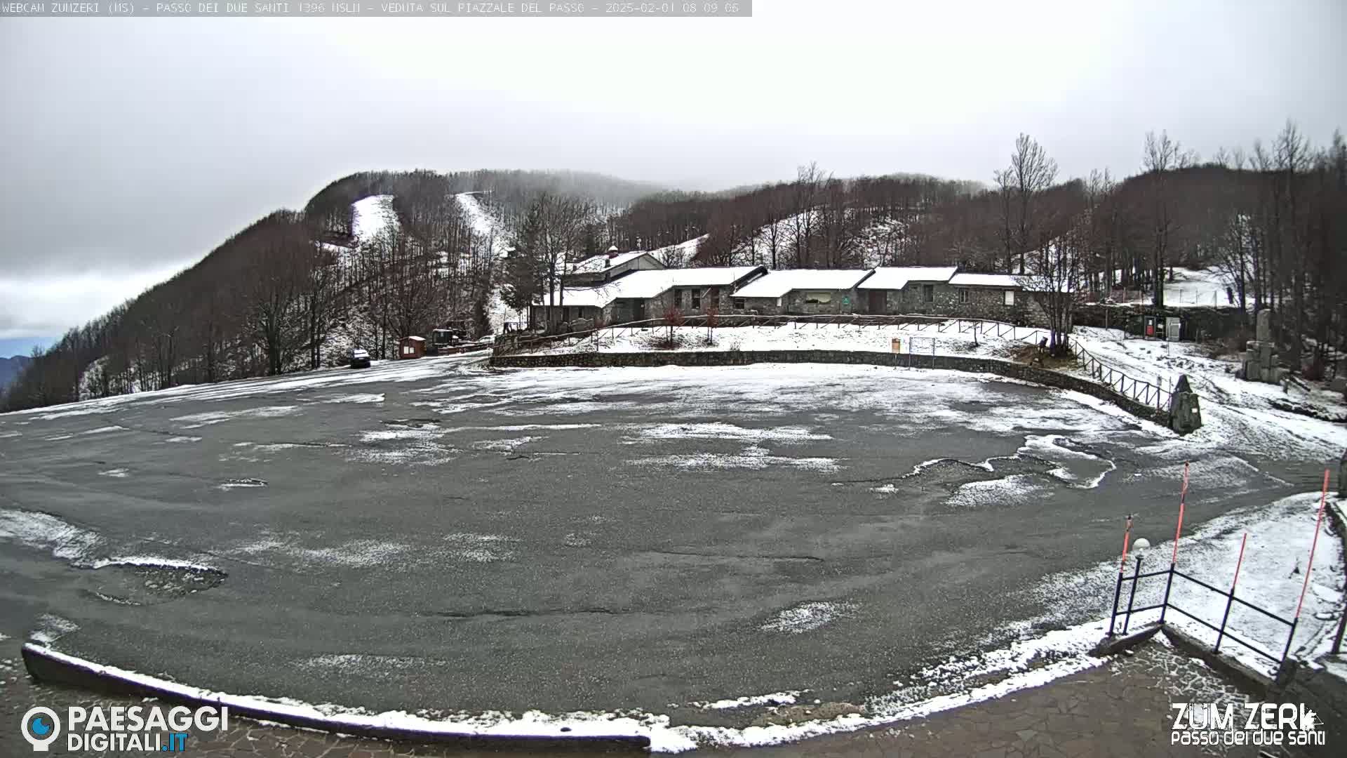 A large, snow-dusted asphalt parking lot sits before a row of stone buildings nestled among snow-covered hills and a ski slope under an overcast sky.