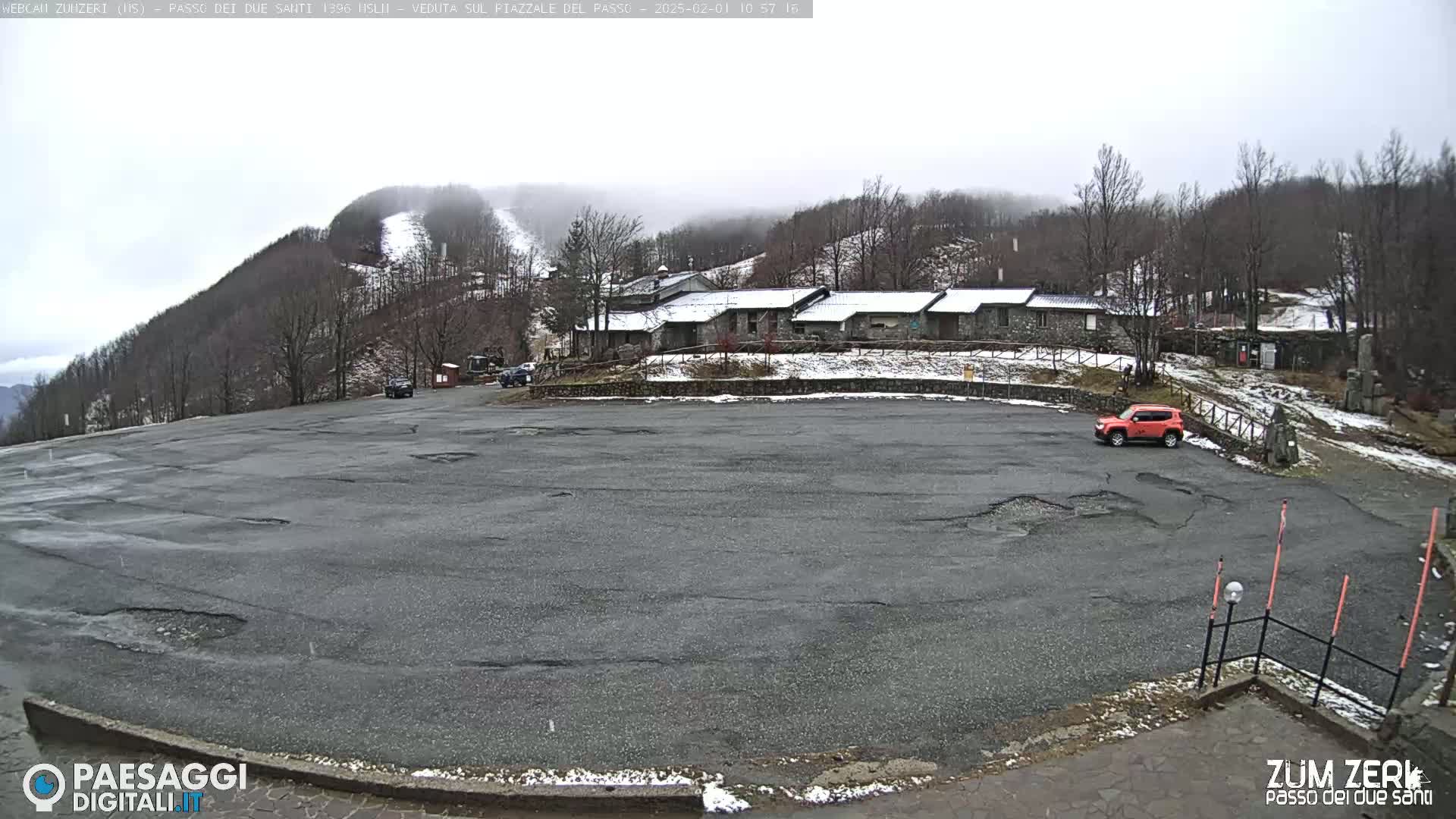 A mostly empty asphalt parking lot sits before a cluster of stone buildings, partially covered in snow, with a snow-dusted hillside and ski slope in the background under an overcast sky.