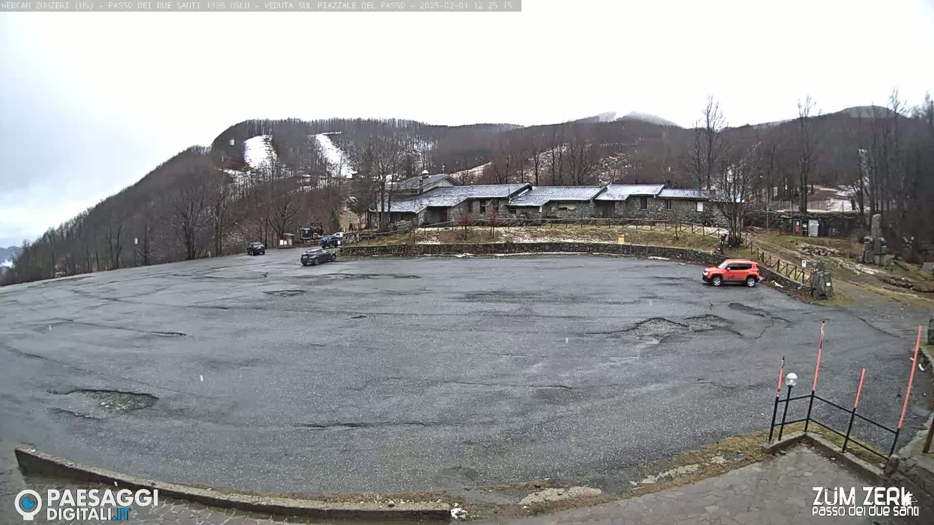 A mostly empty asphalt parking lot sits in front of a stone building complex nestled among snow-dusted hills under an overcast sky.