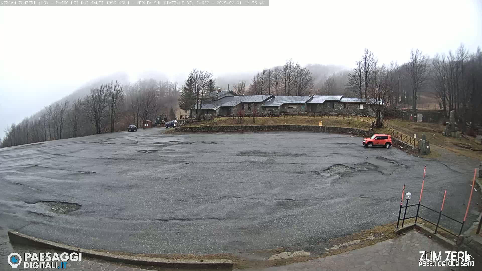 A large, empty asphalt parking lot with potholes is situated in front of a stone building complex, all surrounded by bare trees and hills under an overcast sky.