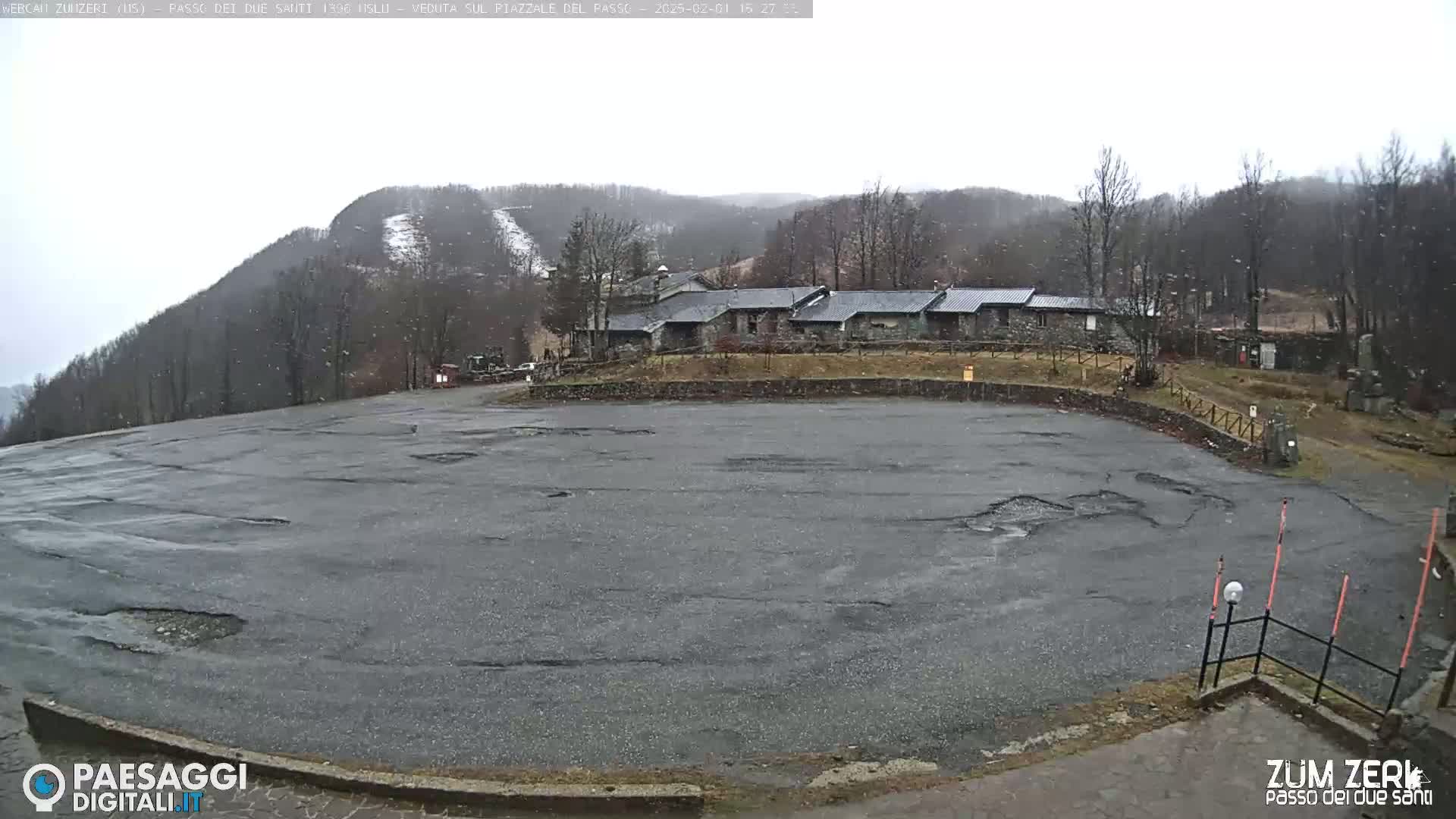 A large, empty asphalt parking lot is seen with light snow falling, in front of a row of buildings nestled against a snow-dusted mountainside.