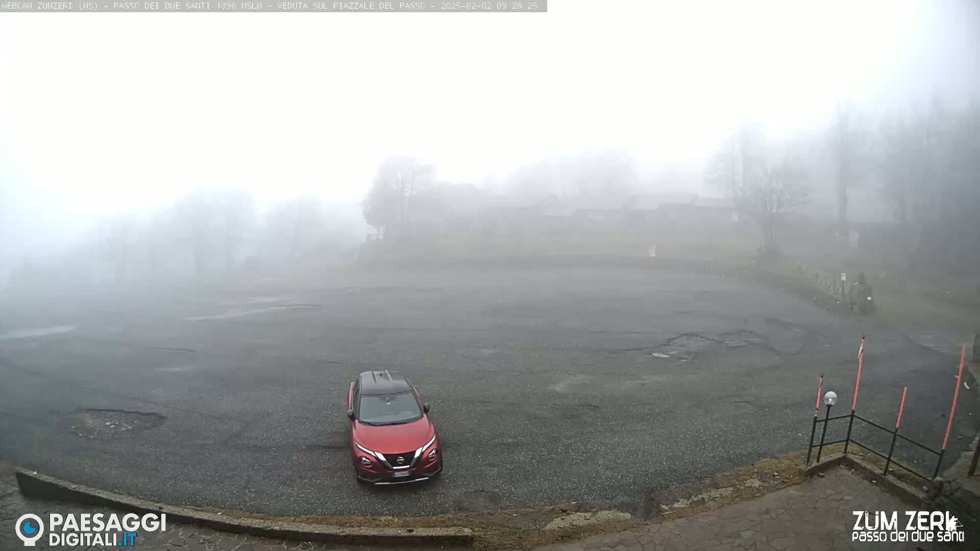 A red SUV is parked in an empty, fog-shrouded parking lot near some trees and buildings.
