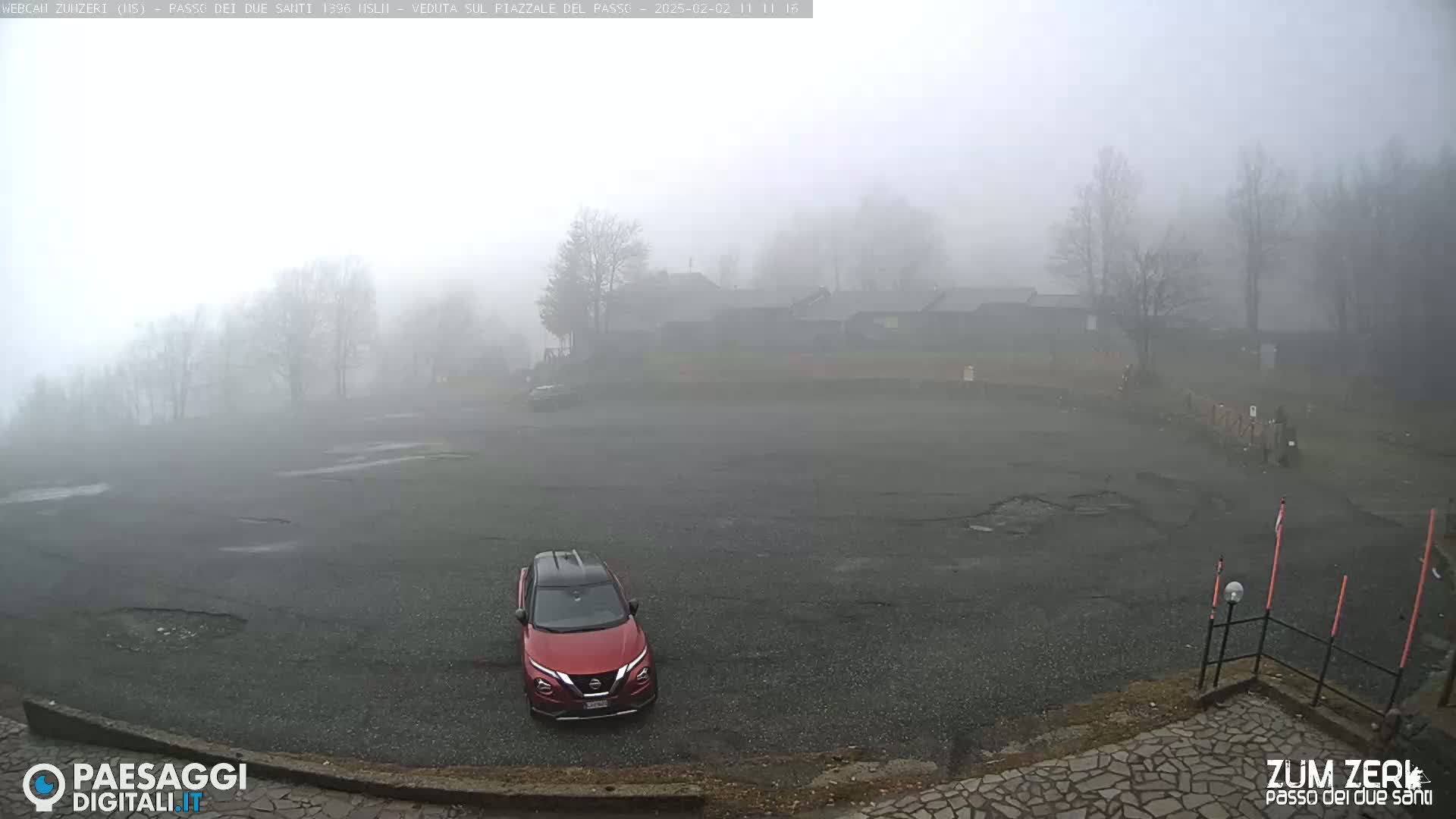 A red SUV is parked in a large, empty asphalt lot shrouded in dense fog, with some buildings and trees barely visible in the background.