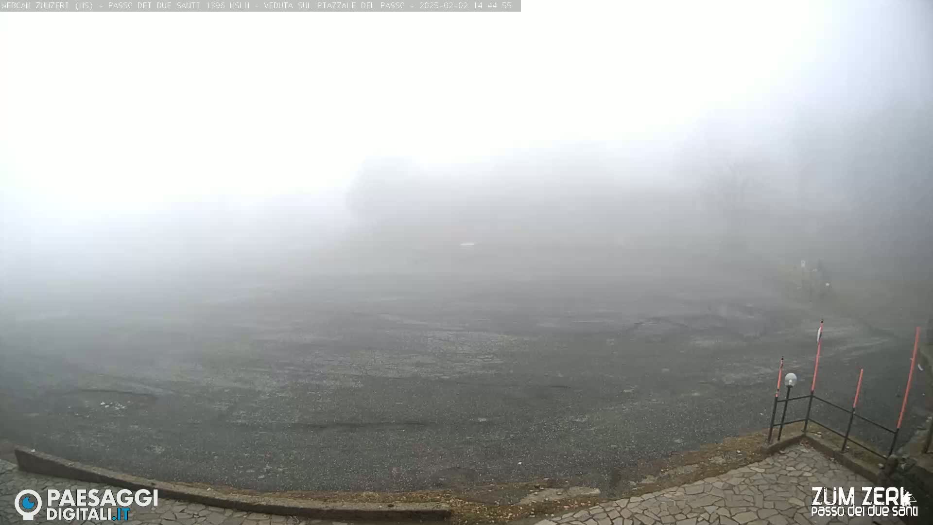 A large, dark-colored paved area is shrouded in thick fog, with indistinct shapes of trees and a railing visible in the lower right corner.