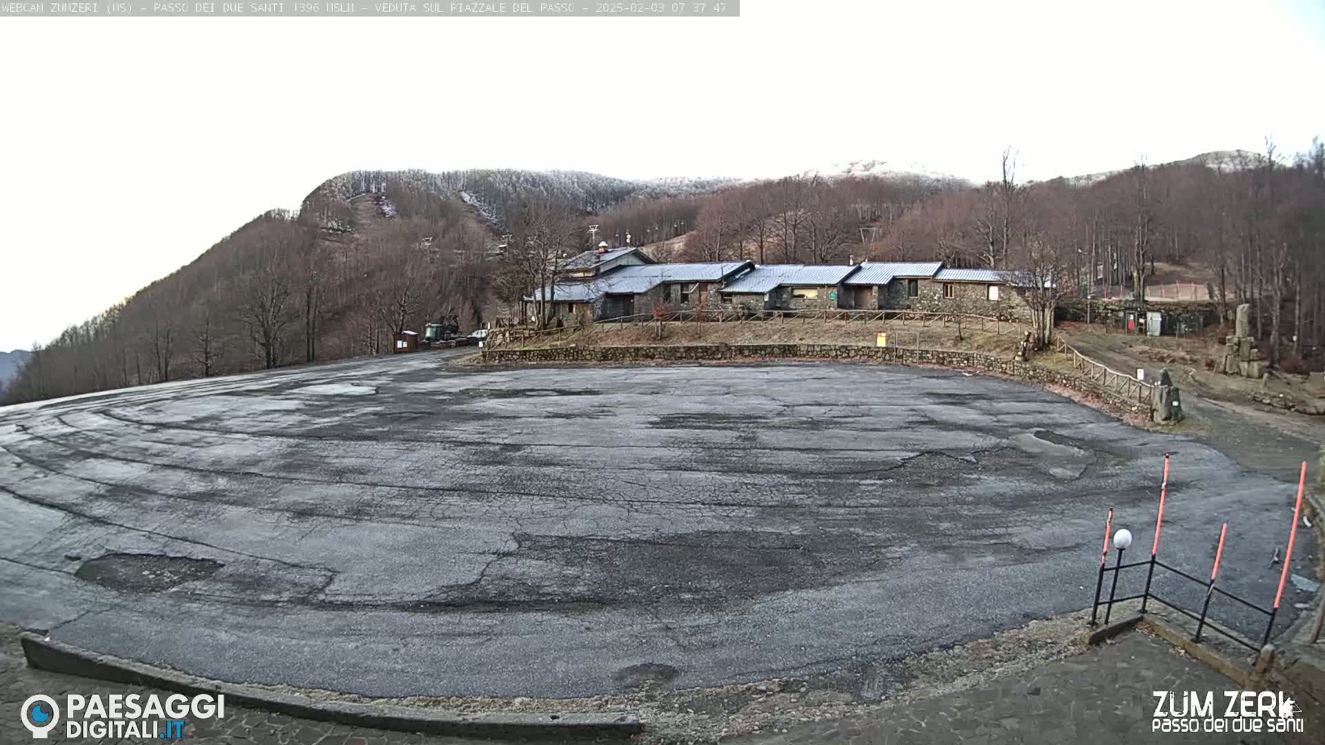 A large, cracked asphalt parking lot sits before a cluster of stone buildings nestled amongst snow-dusted, leafless trees on a mostly cloudy day.