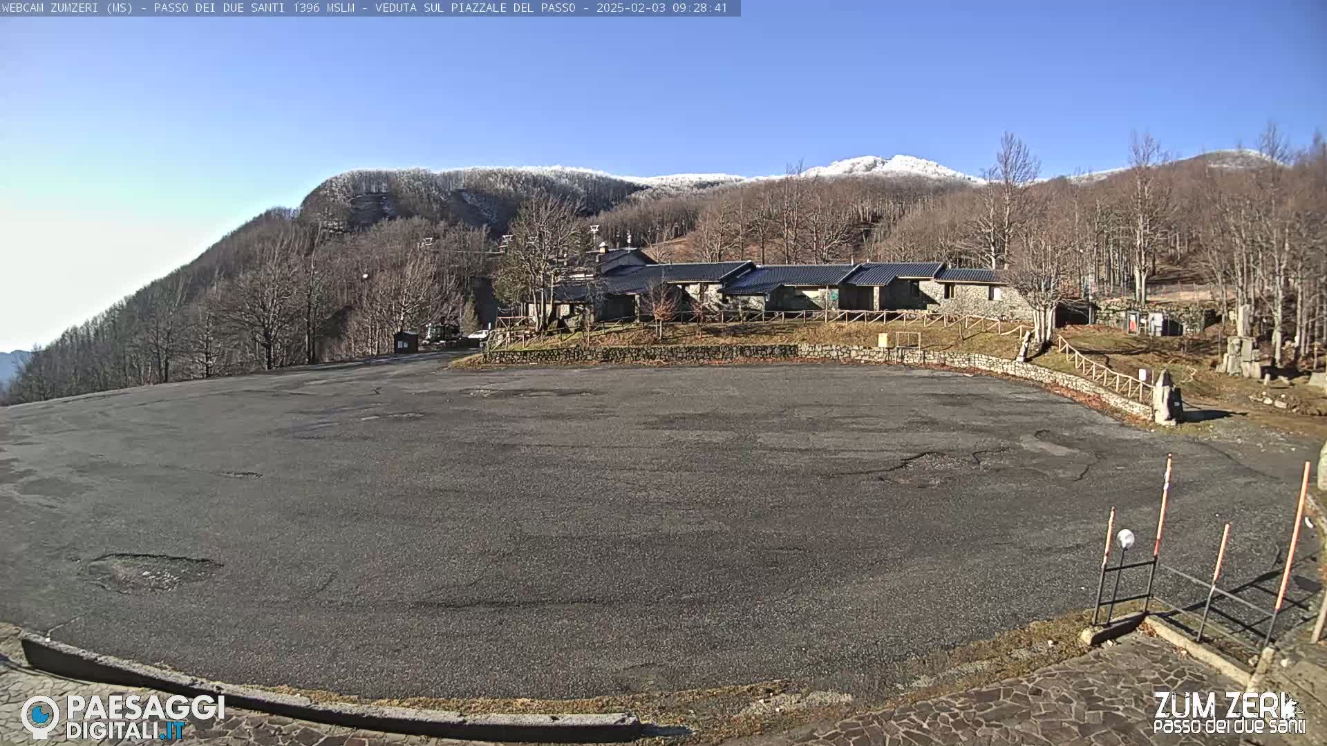 A large, empty asphalt parking lot sits before a cluster of stone buildings nestled in a snow-dusted mountainside under a clear blue sky.