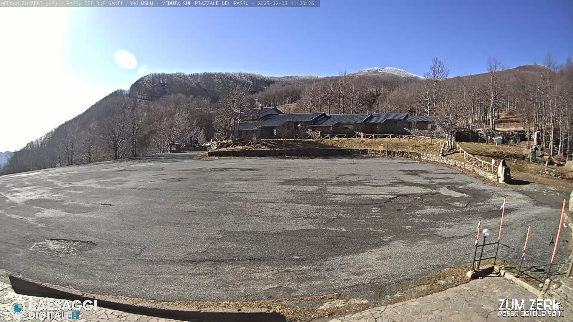 A large, empty asphalt parking lot sits before a row of low buildings, backed by a snow-dusted mountain range under a clear, sunny sky.