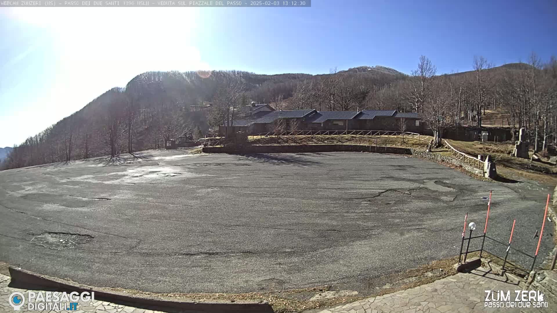 A large, empty asphalt parking lot sits before a stone building nestled in a mountainous area under a bright, sunny sky.