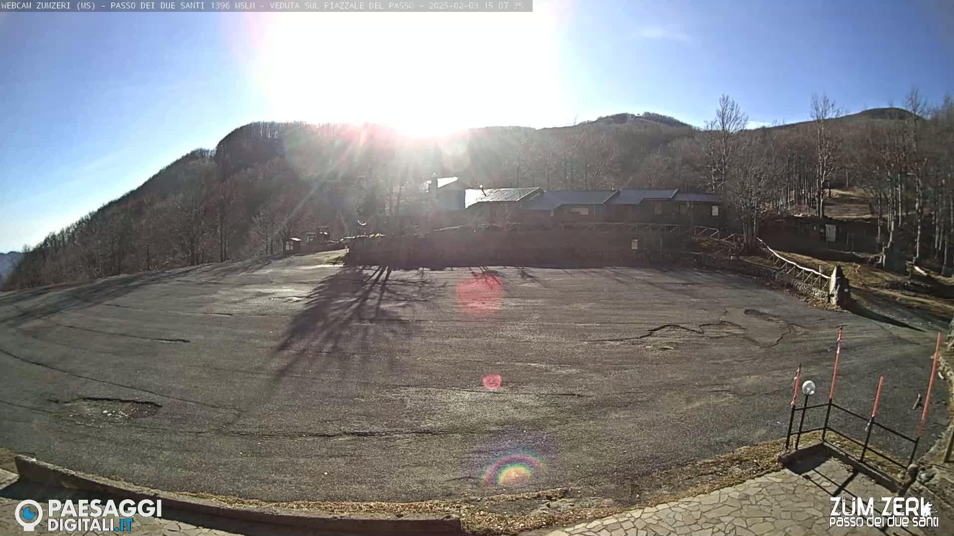 A sunny day reveals an empty asphalt parking lot overlooking a mountainside with leafless trees and a few buildings.