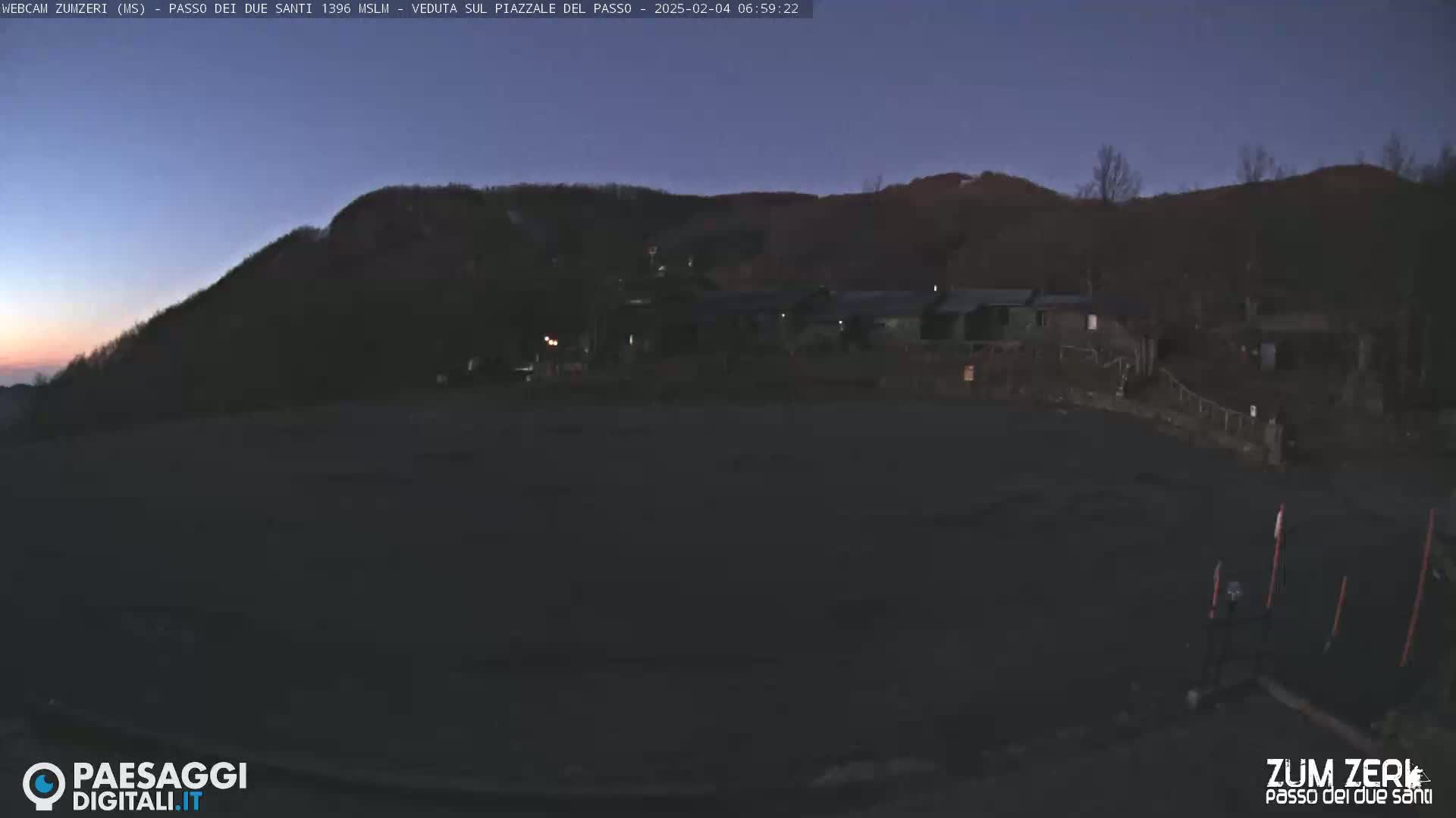 A dark, empty parking lot sits before a line of low buildings at the base of a mountain under a twilight sky.