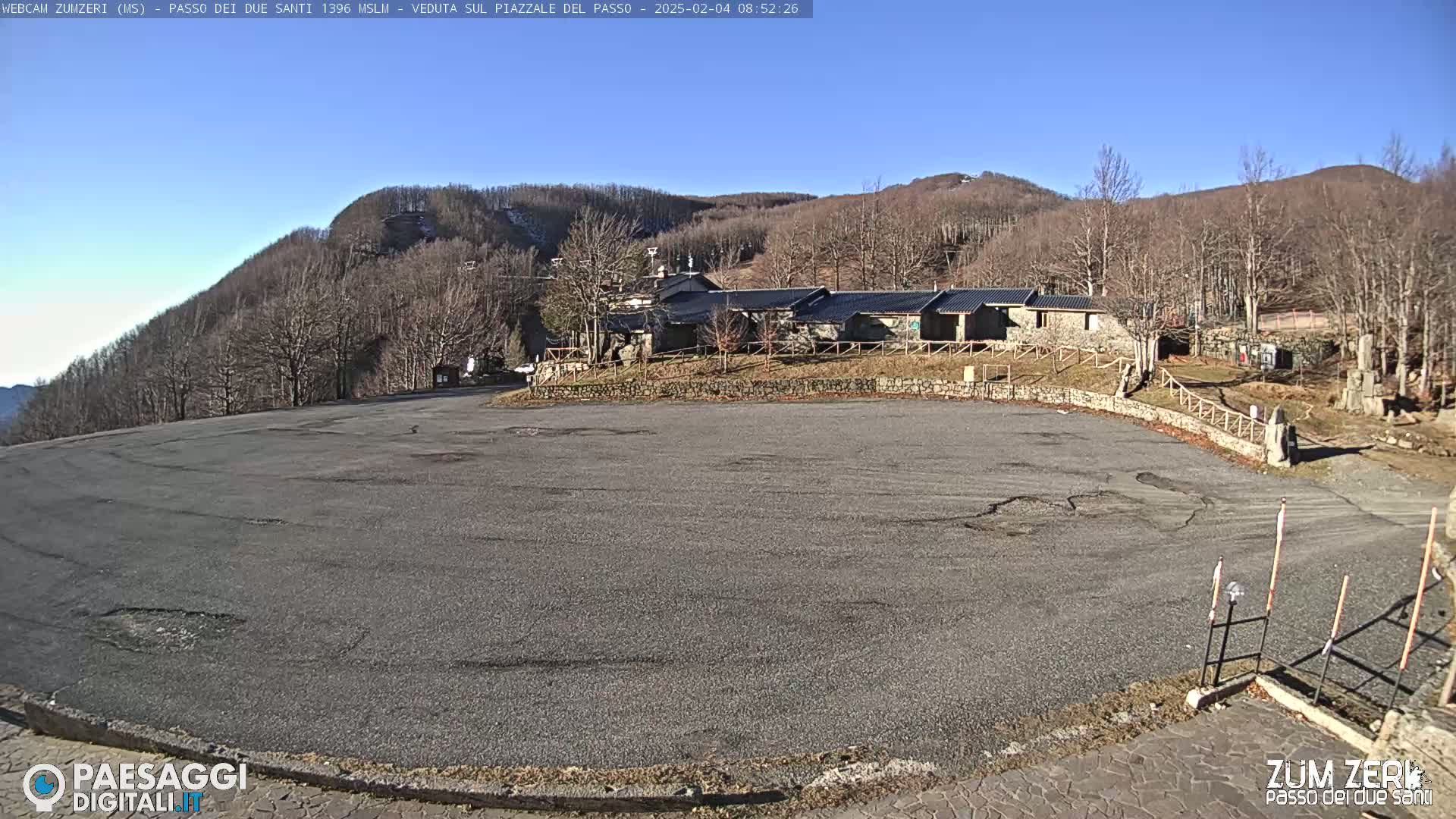 A large, empty asphalt parking lot sits in front of a cluster of stone buildings, all of which are nestled among bare, winter trees on a sunny, clear day.