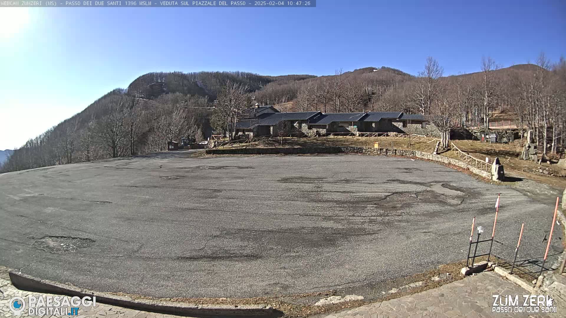 A large, empty asphalt parking lot is situated in front of a row of low buildings, with bare, winter trees and mountains in the background under a clear blue sky.
