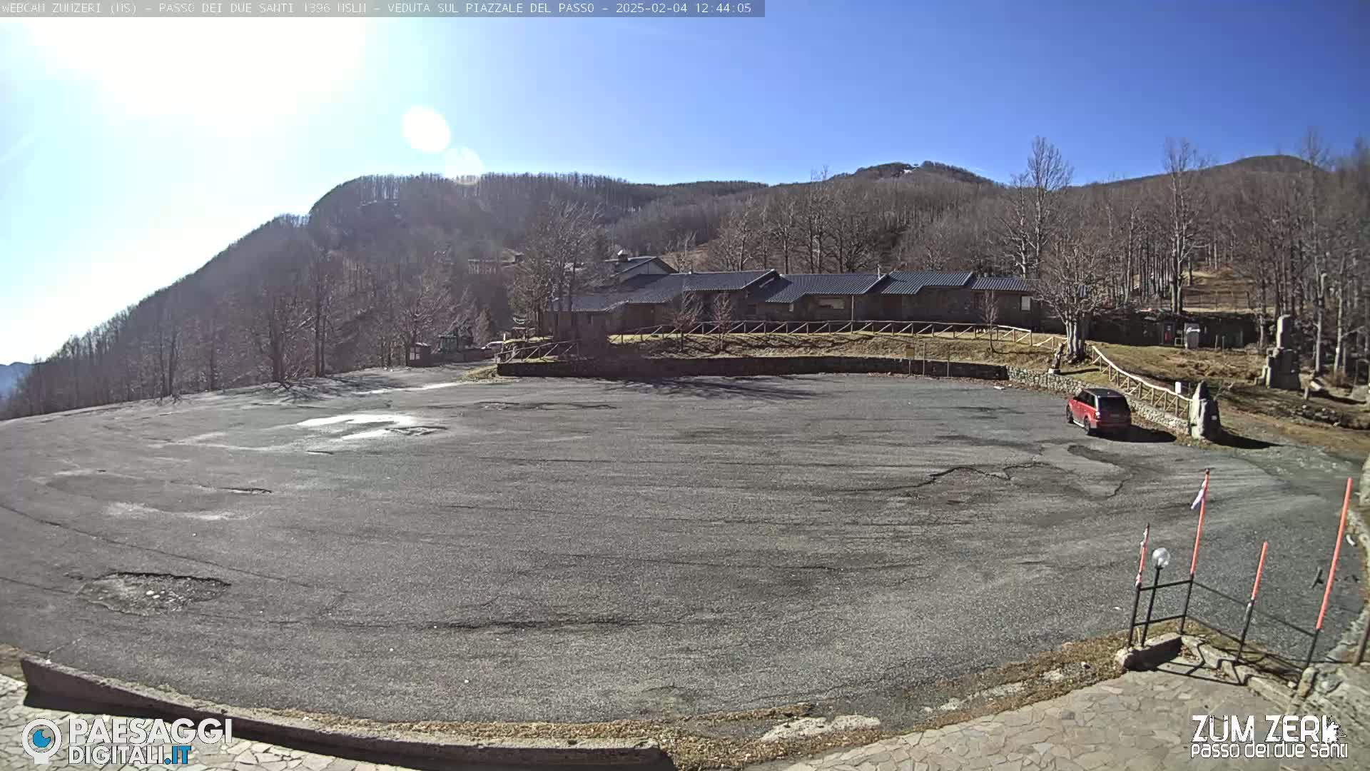 A mostly empty asphalt parking lot sits in front of a low stone building, backed by a hillside of leafless trees under a clear blue sky.