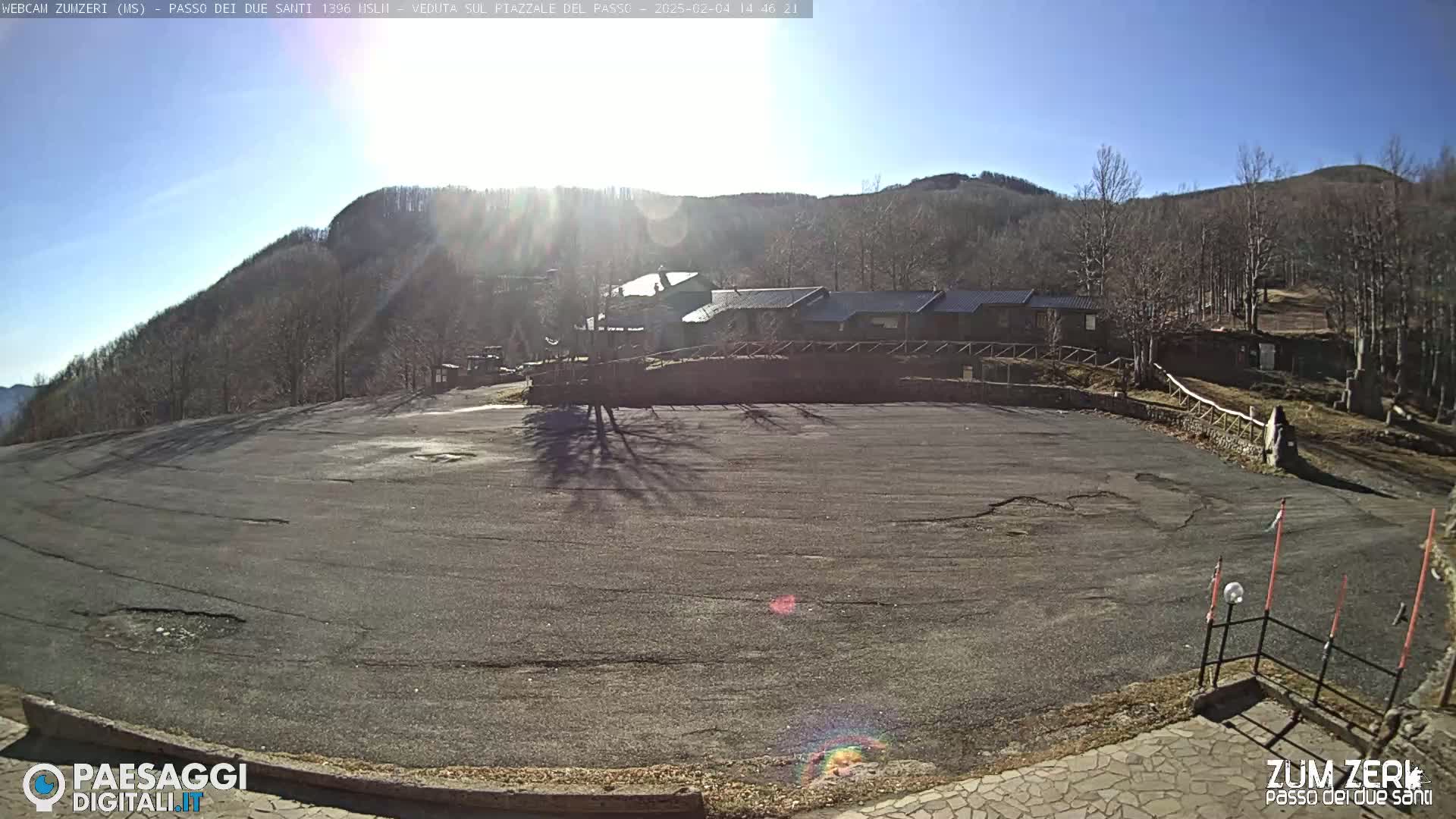 A large, empty asphalt parking lot sits in front of a low building complex nestled among bare, winter trees on a sunny mountainside.