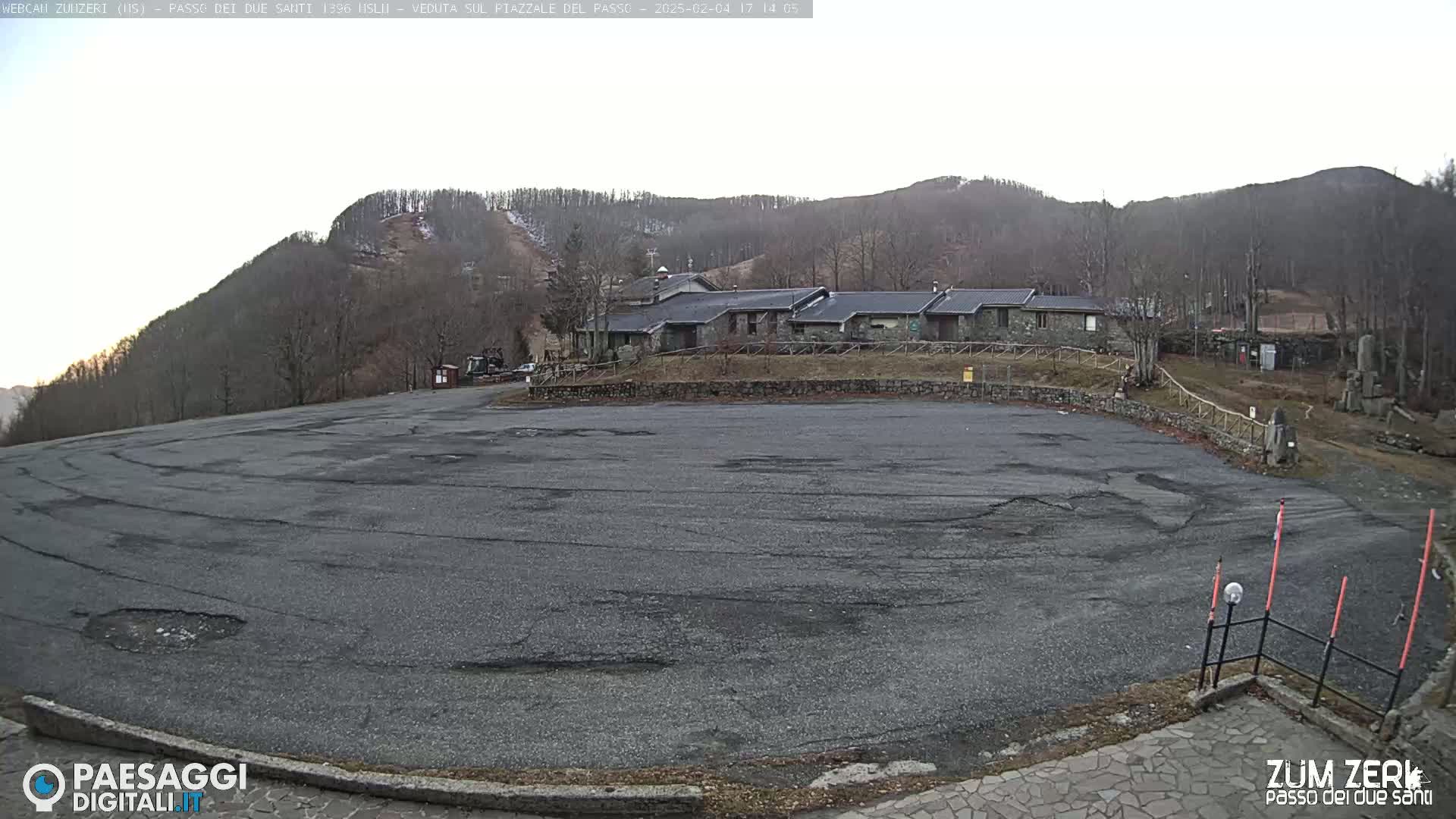 A large, empty asphalt parking lot sits before a stone building complex nestled in a mountainside under an overcast sky.