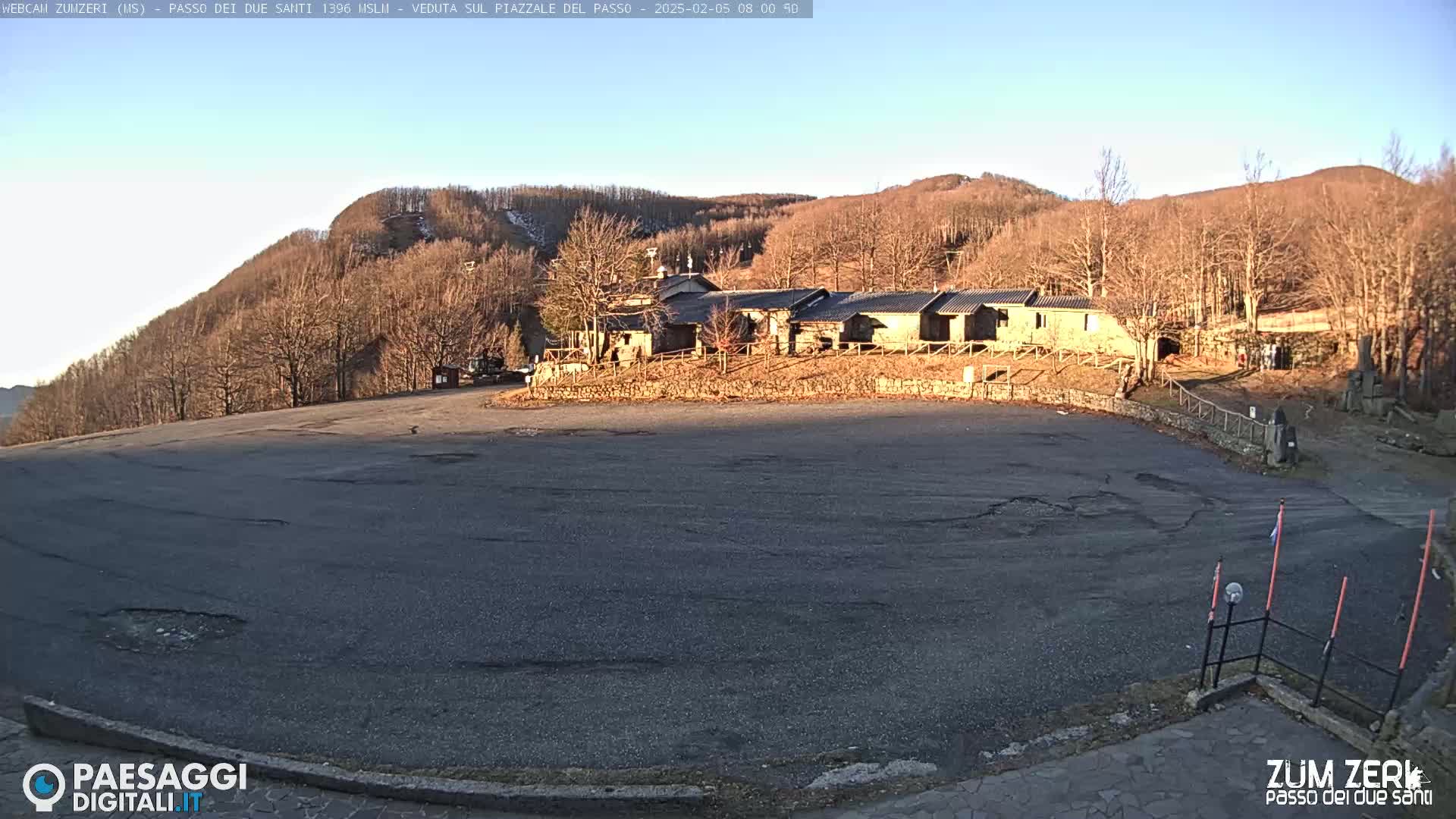 A mostly empty asphalt parking lot sits in front of a stone building complex nestled amongst leafless trees on a sunny day.