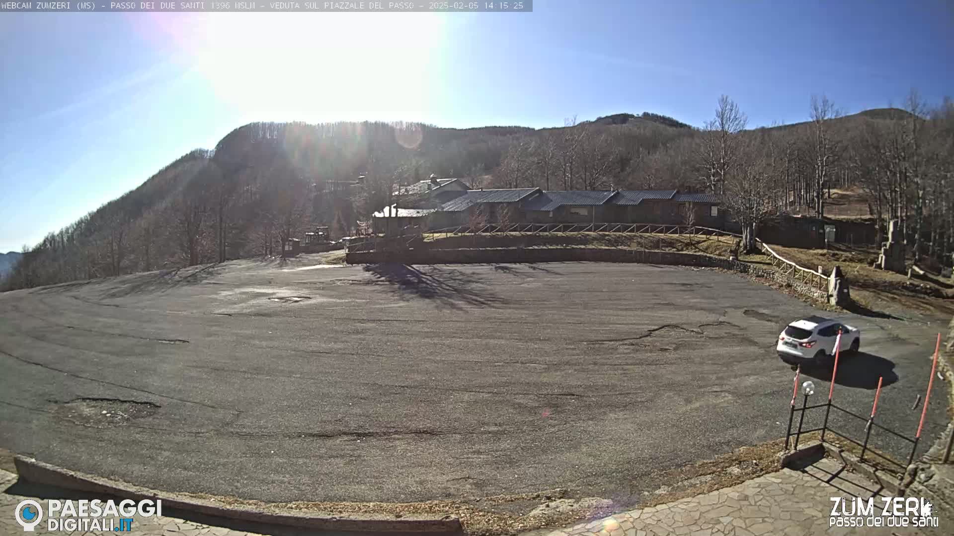 A mostly empty asphalt parking lot is situated in front of a low stone building, with a white SUV parked nearby, all under a bright, sunny sky and against a backdrop of leafless trees and a mountain.