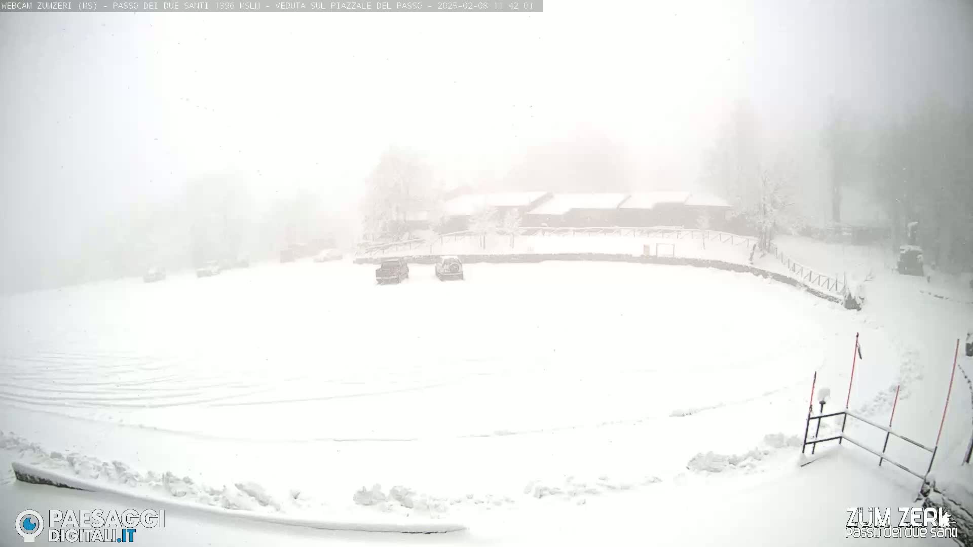 A snow-covered plaza with a few vehicles and buildings in the background during a heavy snowstorm.