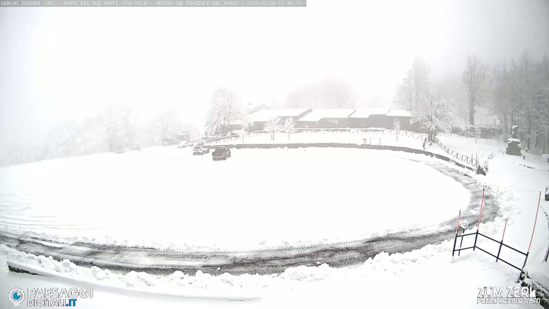 A snow-covered parking lot and roadway curve around a snow-covered building complex in heavy snow.
