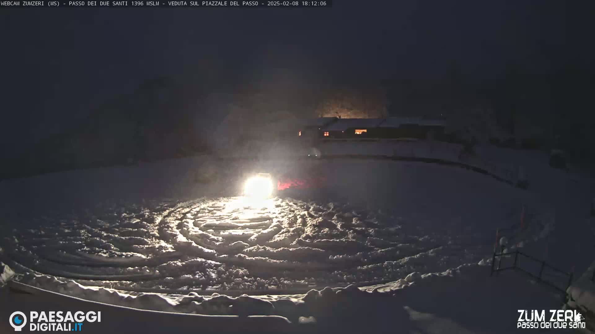 A snow-covered area at night is illuminated by the headlights of a vehicle making circular patterns in the snow, with a building visible in the background.