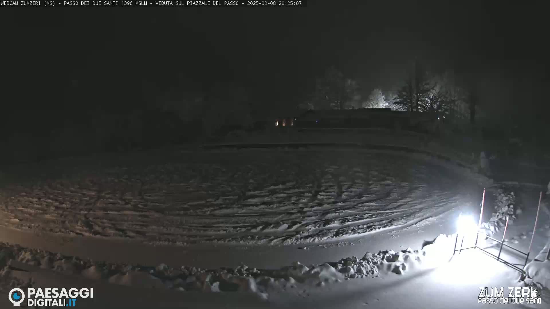 A snow-covered plaza is illuminated at night by a single light source, with dark trees visible in the background under a foggy sky.