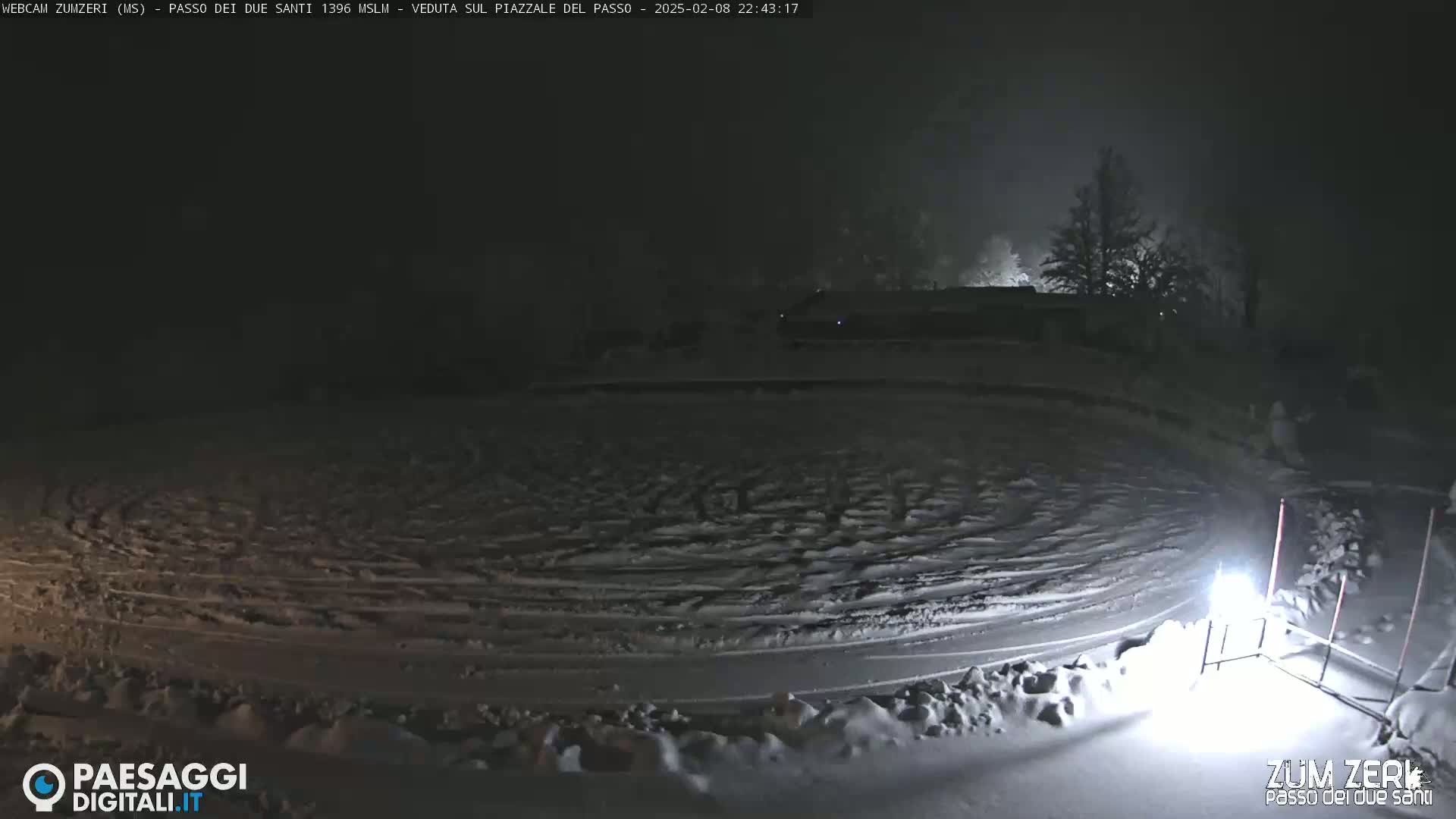A snow-covered circular area with tire tracks is visible at night under foggy conditions, with dimly lit trees and buildings in the background.