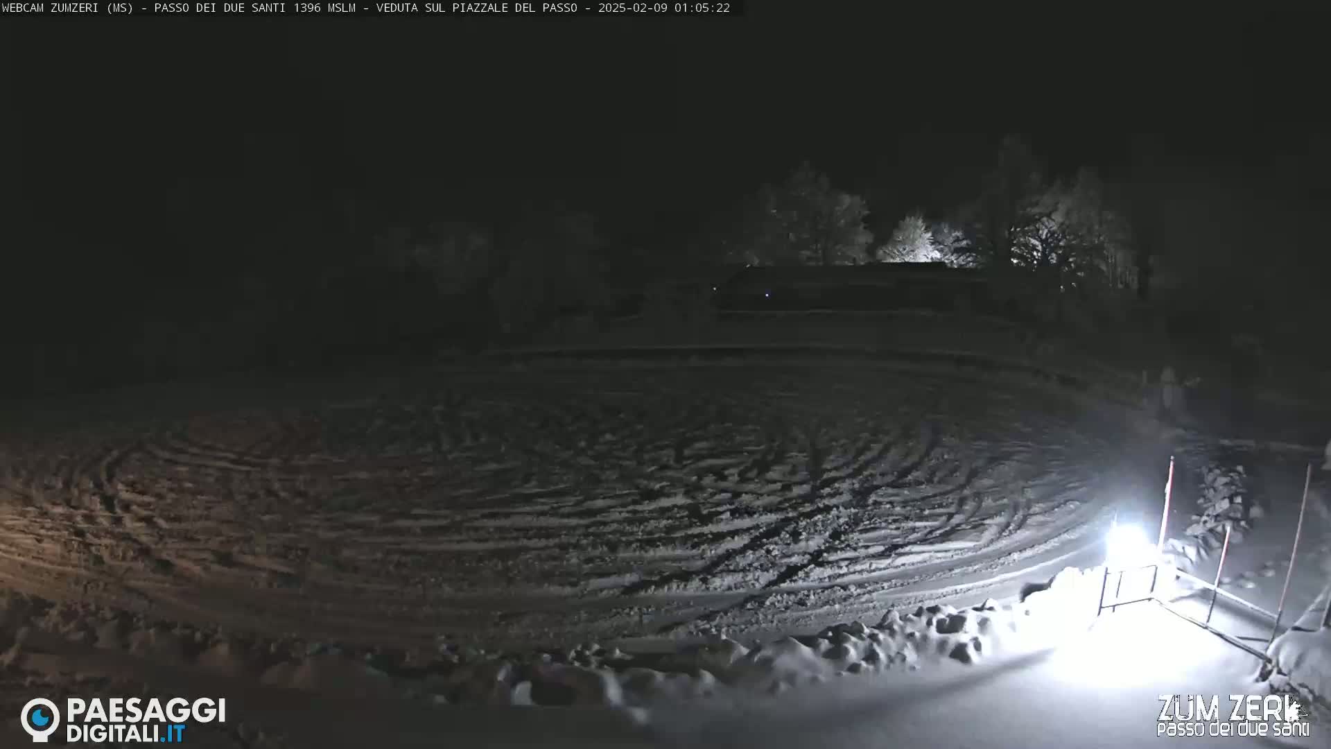 A snow-covered, circular area with tire tracks is visible at night, under dark skies, surrounded by sparsely-lit trees.