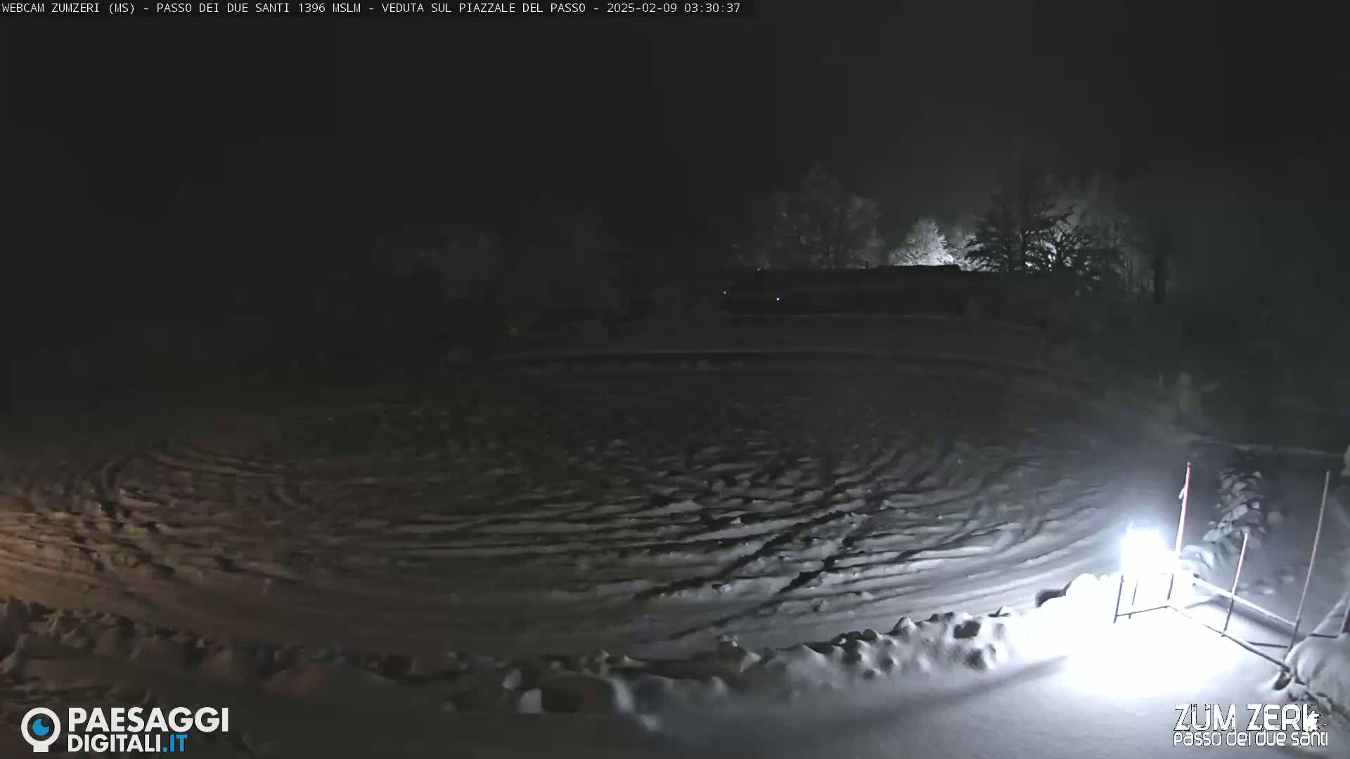 A snow-covered, dark open area with tire tracks and dimly lit trees in the background under a nighttime sky.