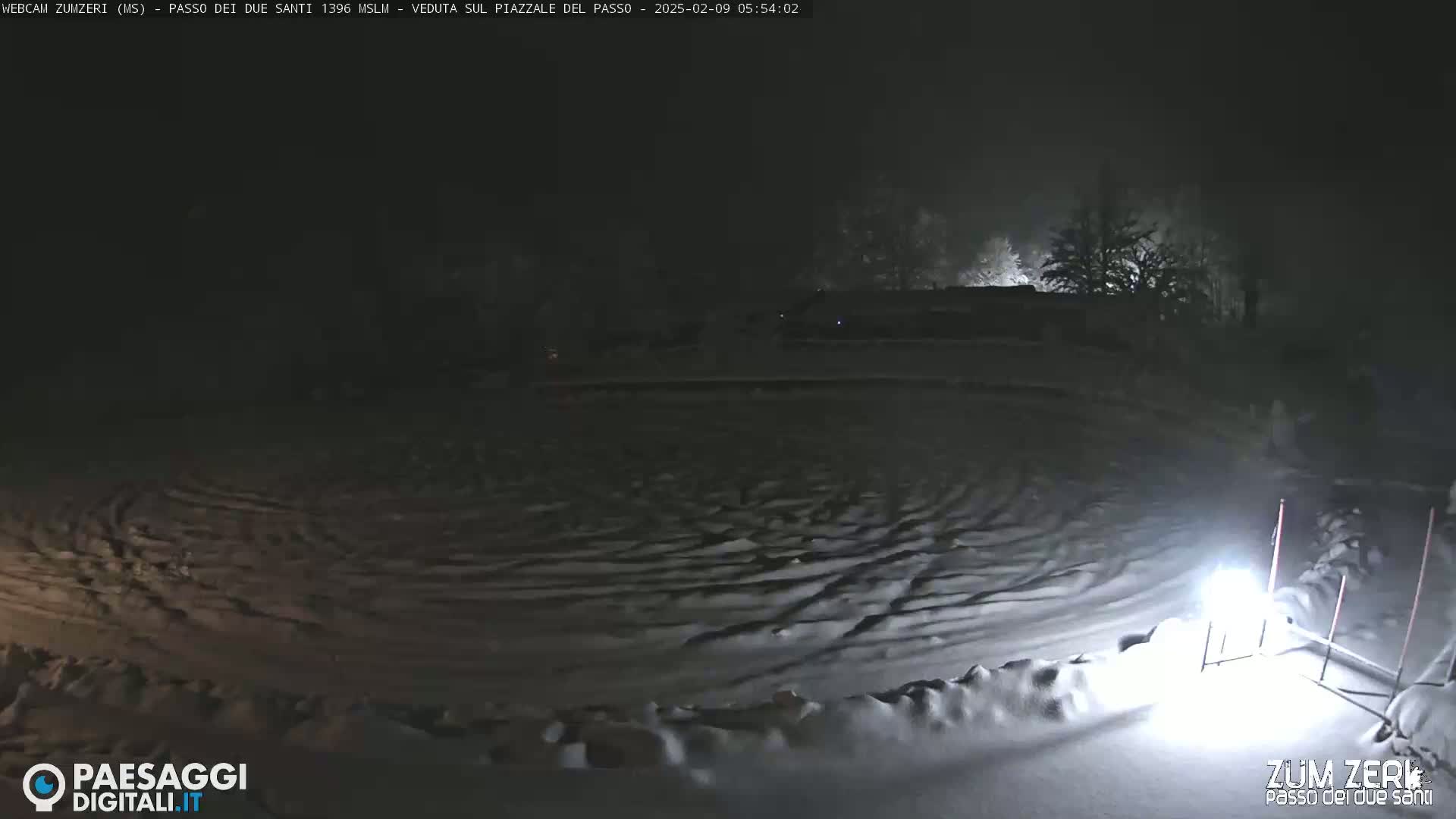 A snow-covered, dark area with tire tracks is illuminated by a bright light at night.