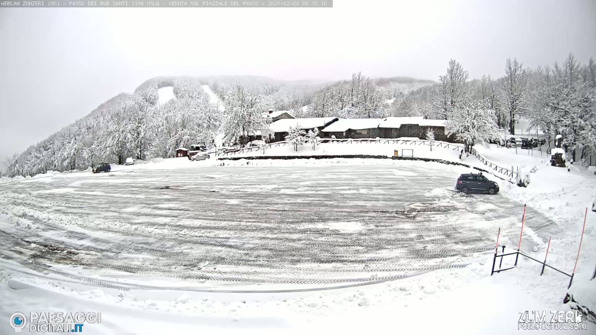 A snow-covered parking lot and building complex are seen on a mountainside under an overcast sky.