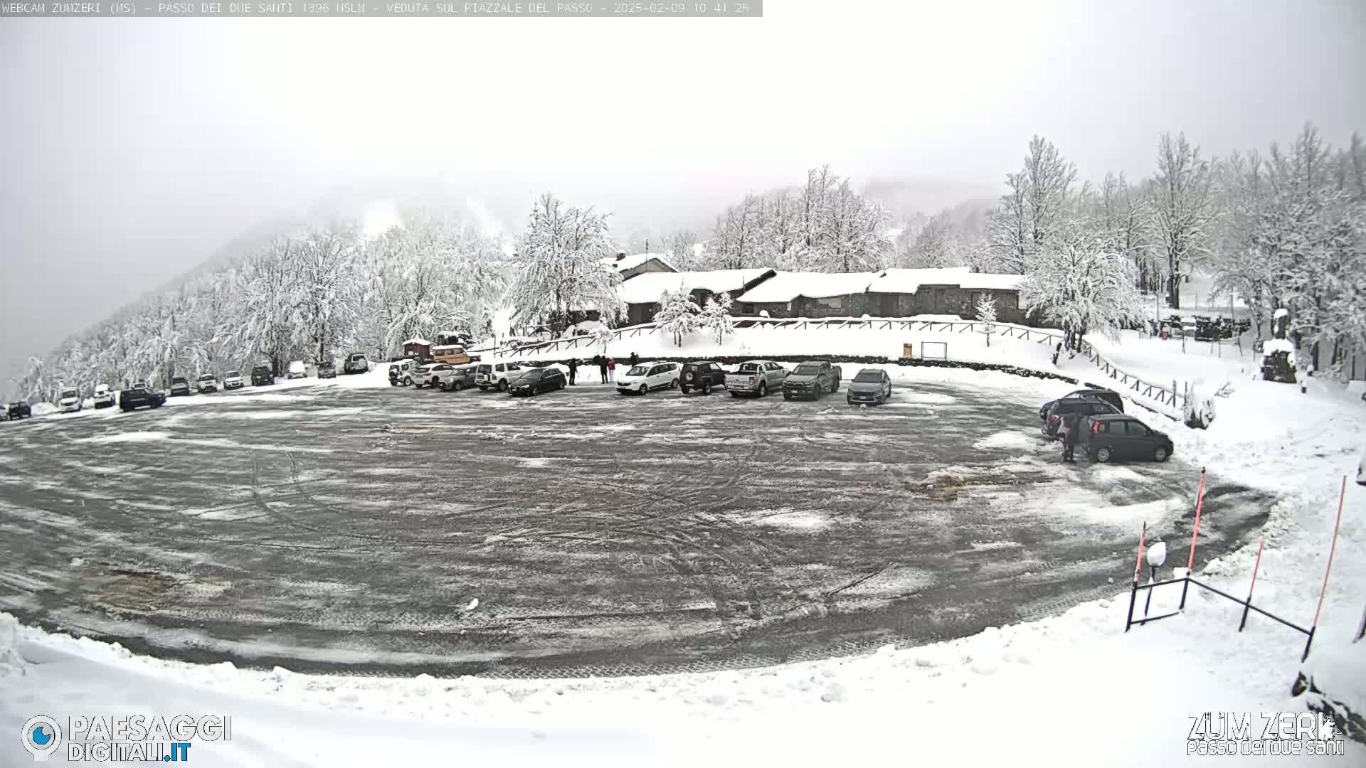 A snow-covered parking lot with several cars and a building in the background under a cloudy sky.