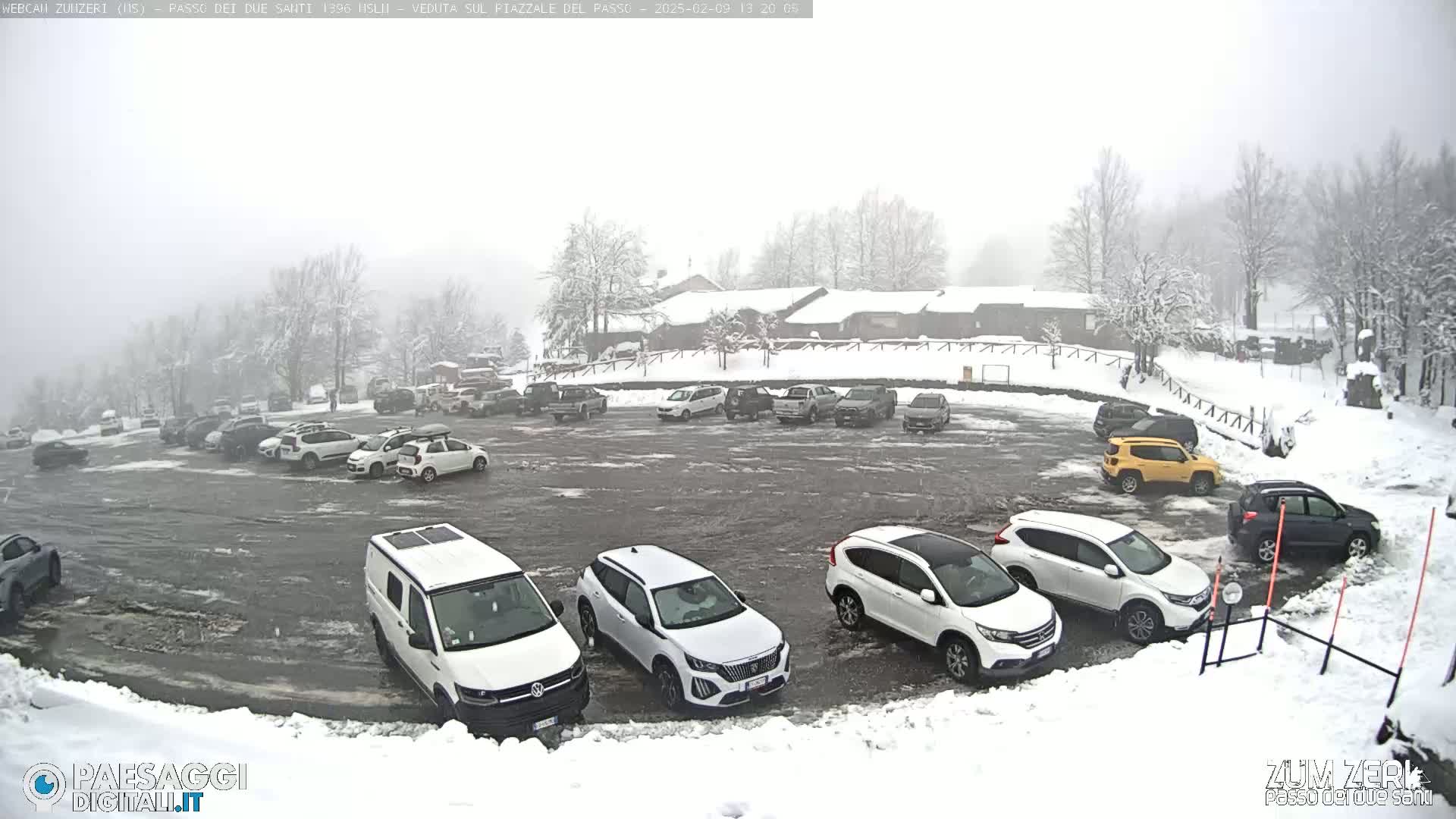 A snow-covered parking lot with numerous cars parked, surrounded by snow-covered trees and a building in the background during a snowfall.