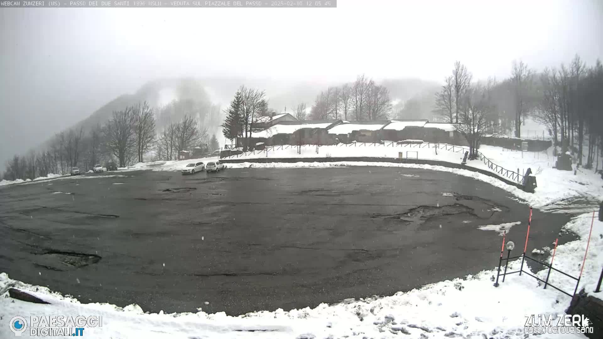A snow-covered parking lot with a few cars and a building in the background, all under a foggy, snowy sky.
