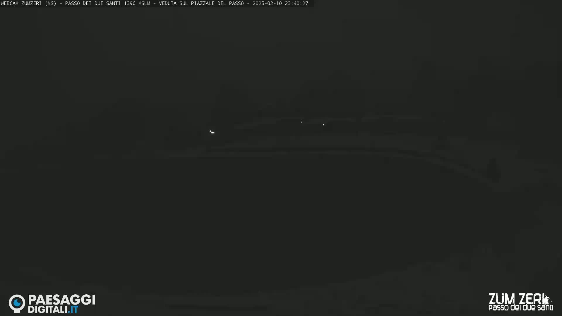 A dark, nighttime view of a dimly lit, snow-covered area with a few faint lights visible in the distance.
