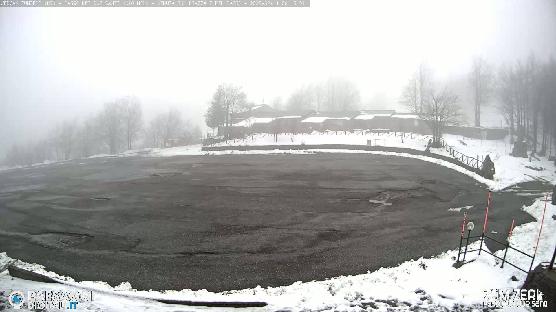 A foggy, snowy parking lot sits before a cluster of buildings nestled among bare trees.