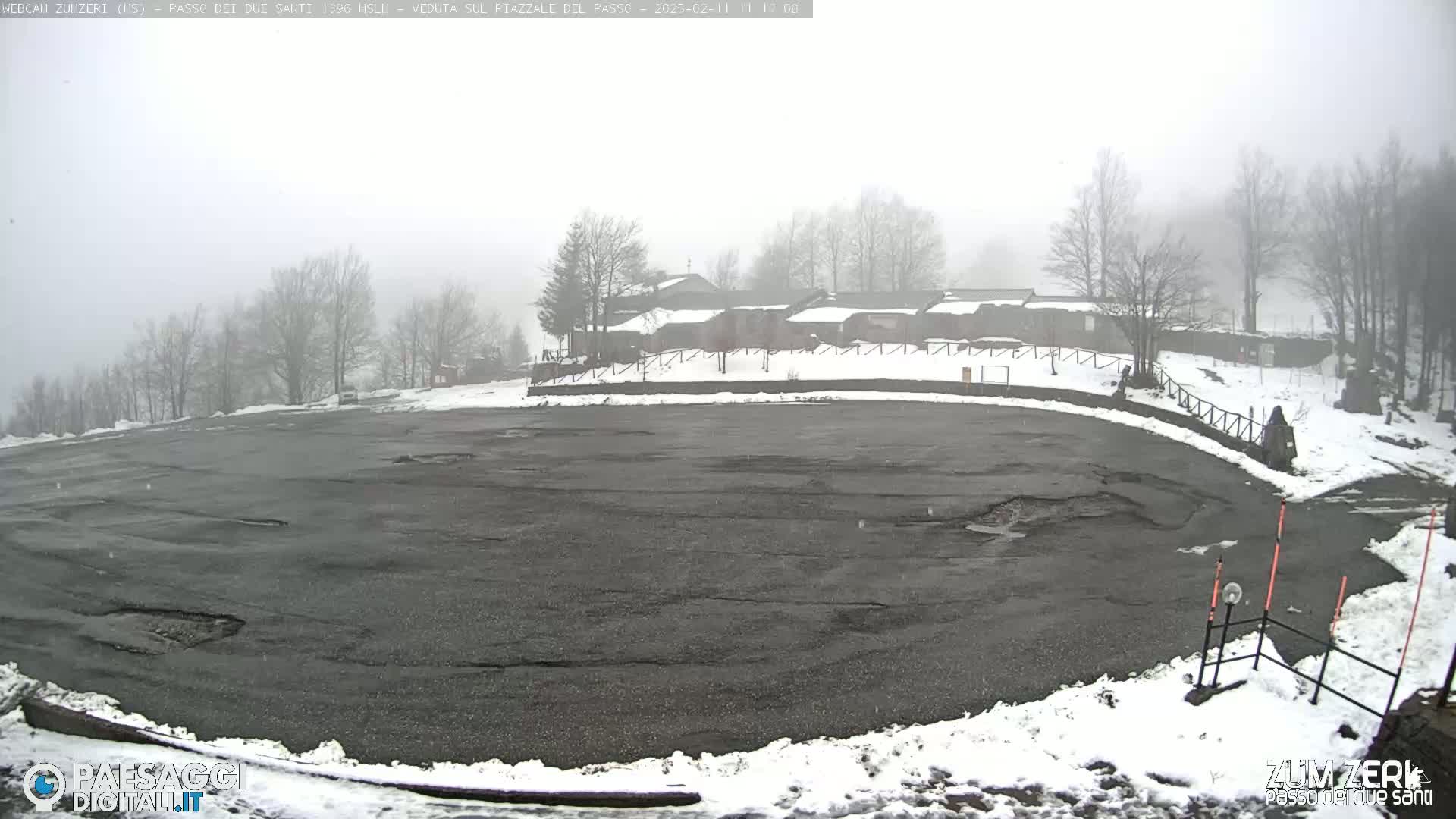 A large, mostly empty asphalt parking lot is partially covered in snow, with a foggy atmosphere and a cluster of buildings visible in the background.