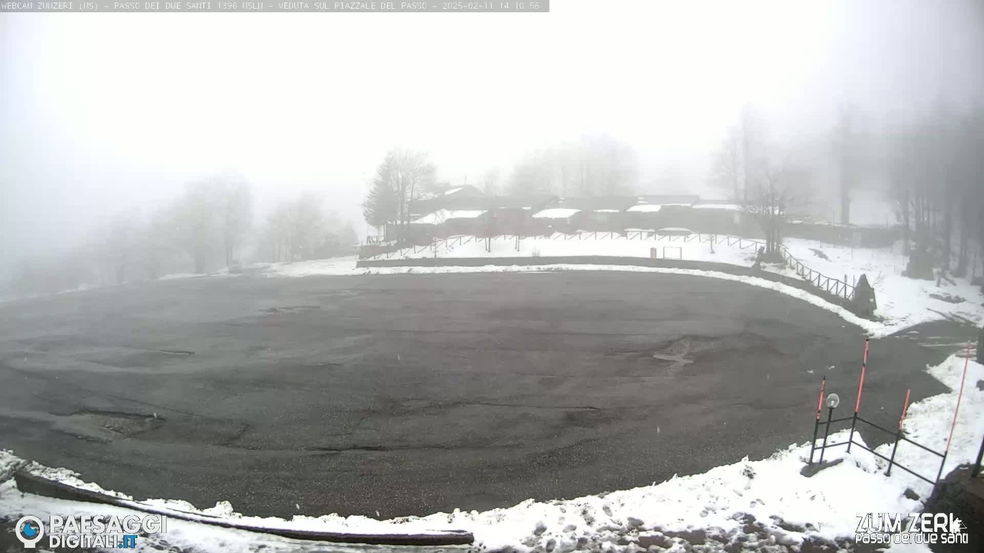 A foggy, snowy scene shows an empty asphalt parking lot next to a row of buildings partially obscured by fog, with some snow on the ground.