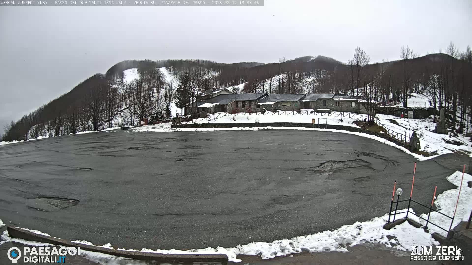 A large, empty asphalt parking lot is situated below a cluster of stone buildings, with snow on the ground and a lightly snow-covered, tree-covered hillside in the background under an overcast sky.