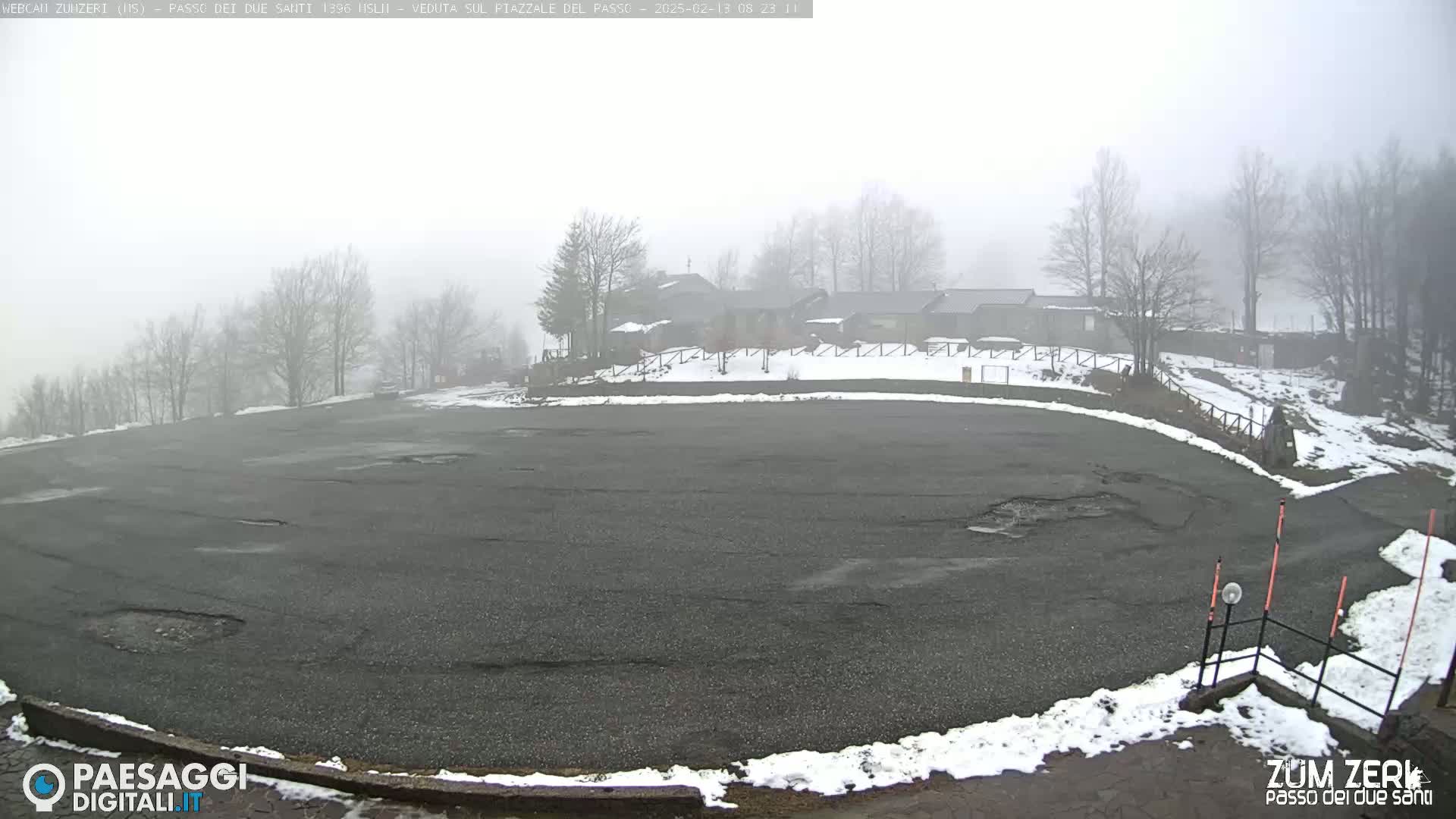 A foggy, snowy parking lot with a few buildings in the background and bare trees surrounding it.