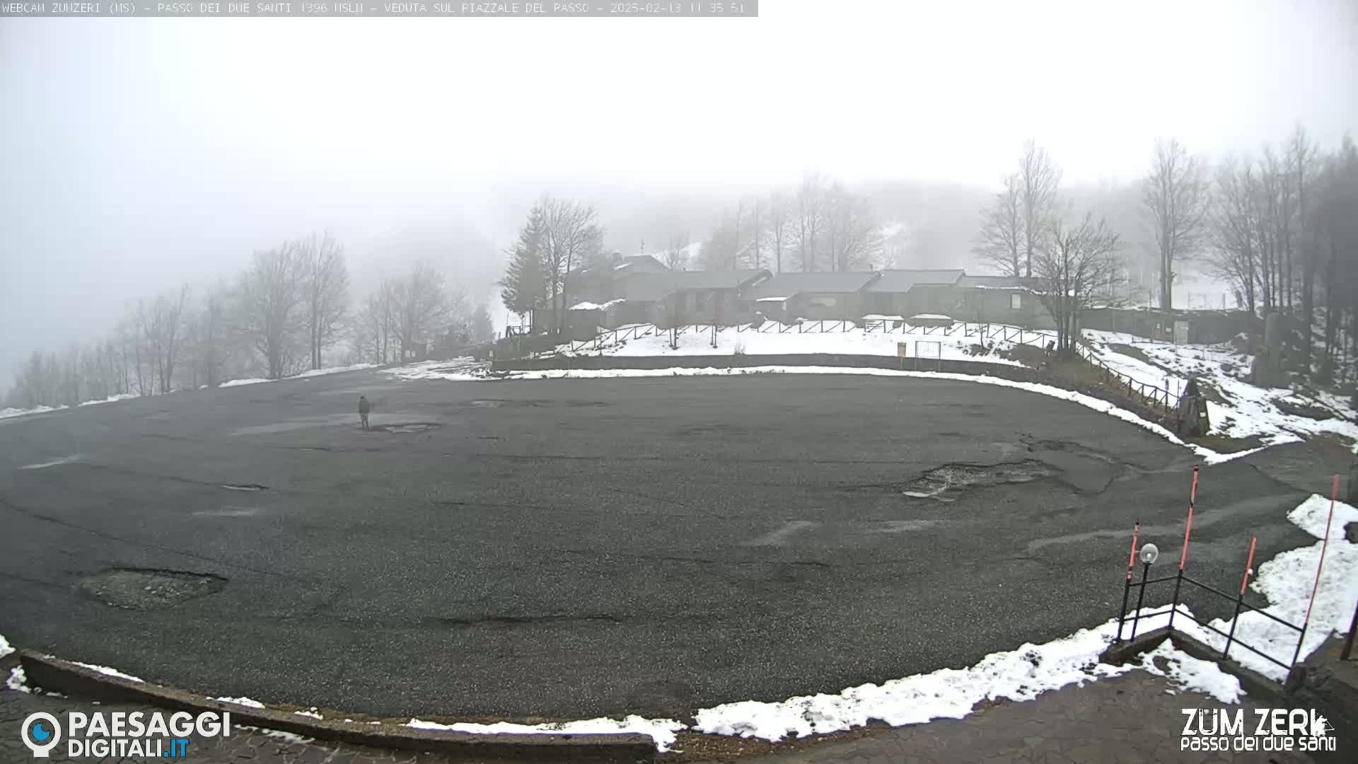 A large, mostly empty asphalt parking lot with some patches of snow and a few bare trees is visible on a foggy day, with a building complex in the background.