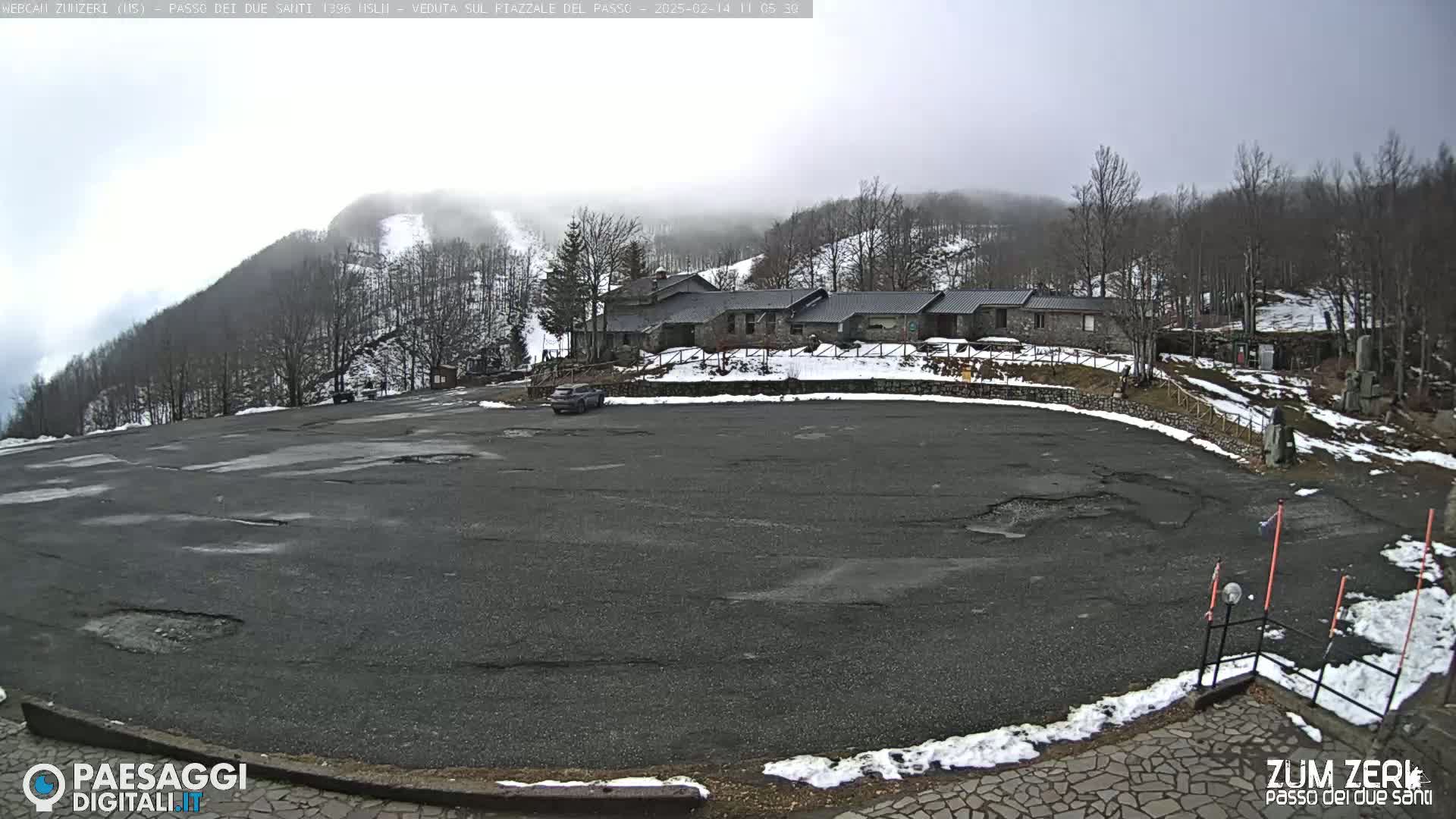 A mostly empty asphalt parking lot is situated in front of a stone building complex, with snow on the ground and a hazy, overcast sky in the background.