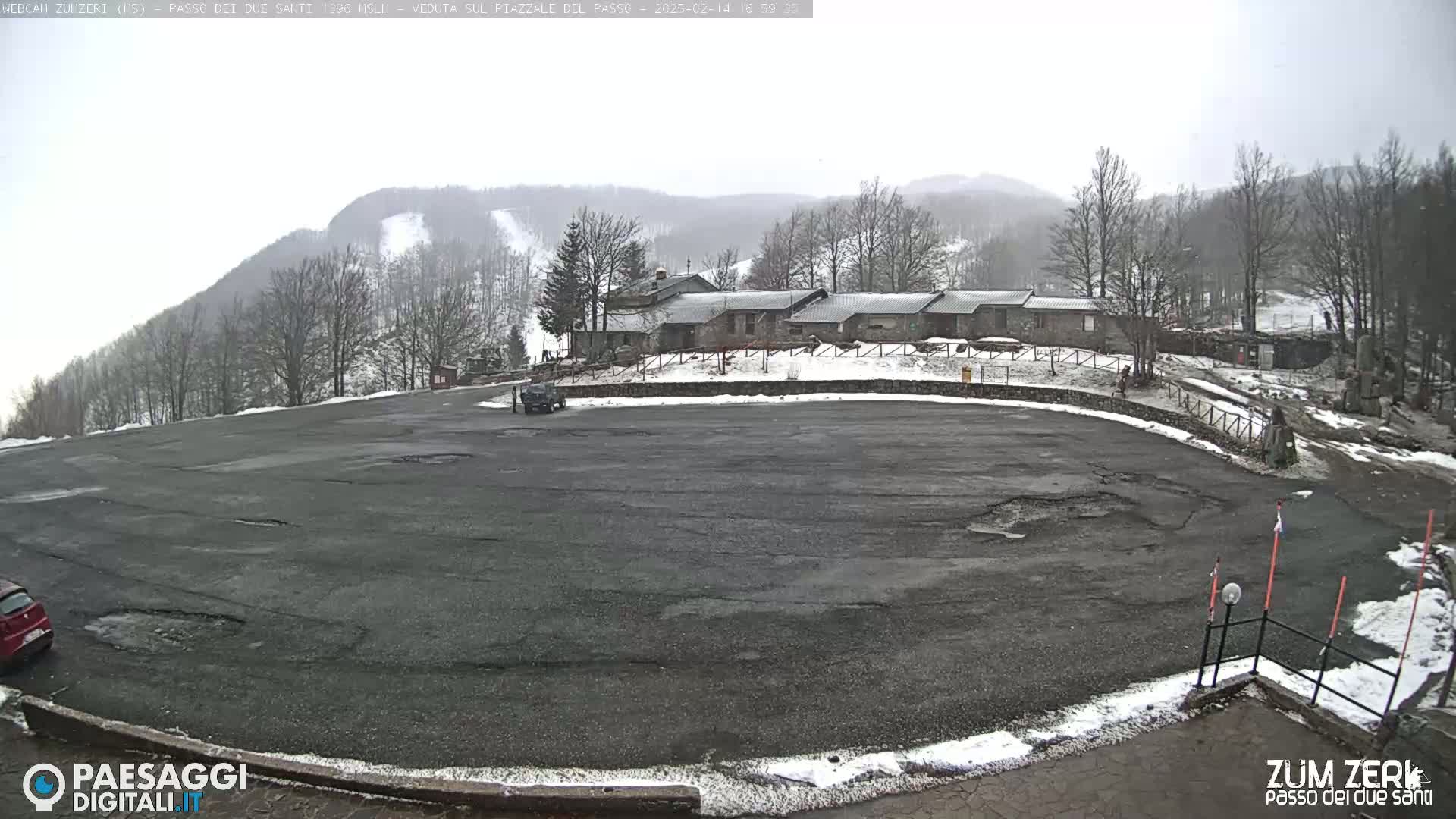 A mostly empty asphalt parking lot is situated in front of a stone building complex, with snow-covered hills and a ski slope visible in the background under an overcast sky.