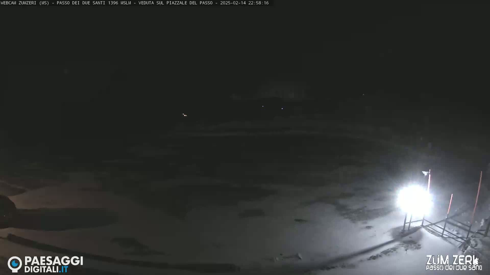 A nighttime, snow-covered scene is illuminated by a bright light source, with faint distant lights visible in the darkness.
