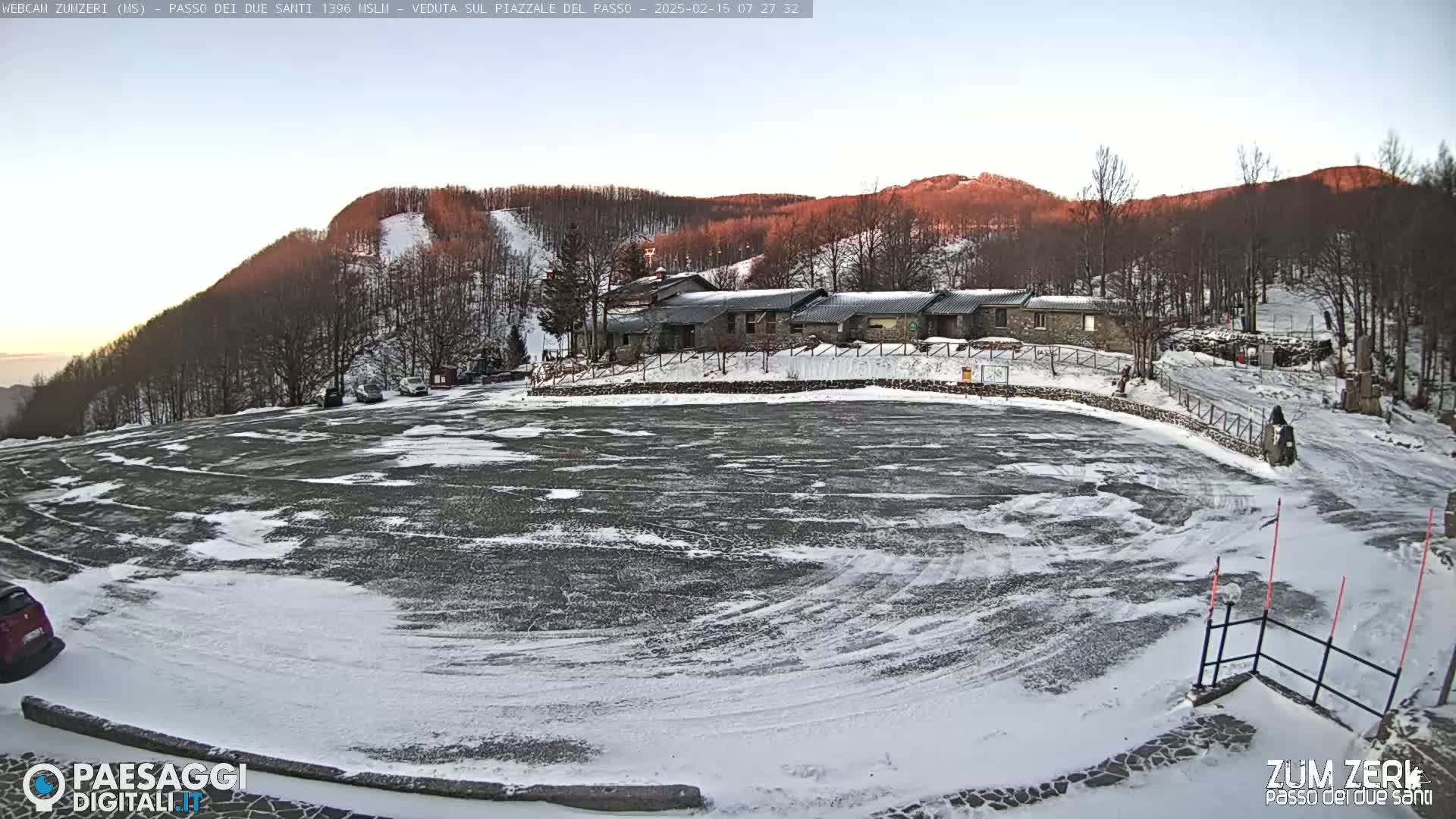 A mostly empty snow-covered parking lot sits before a stone building complex nestled in a snow-dusted mountainside under a clear, pale sunrise sky.