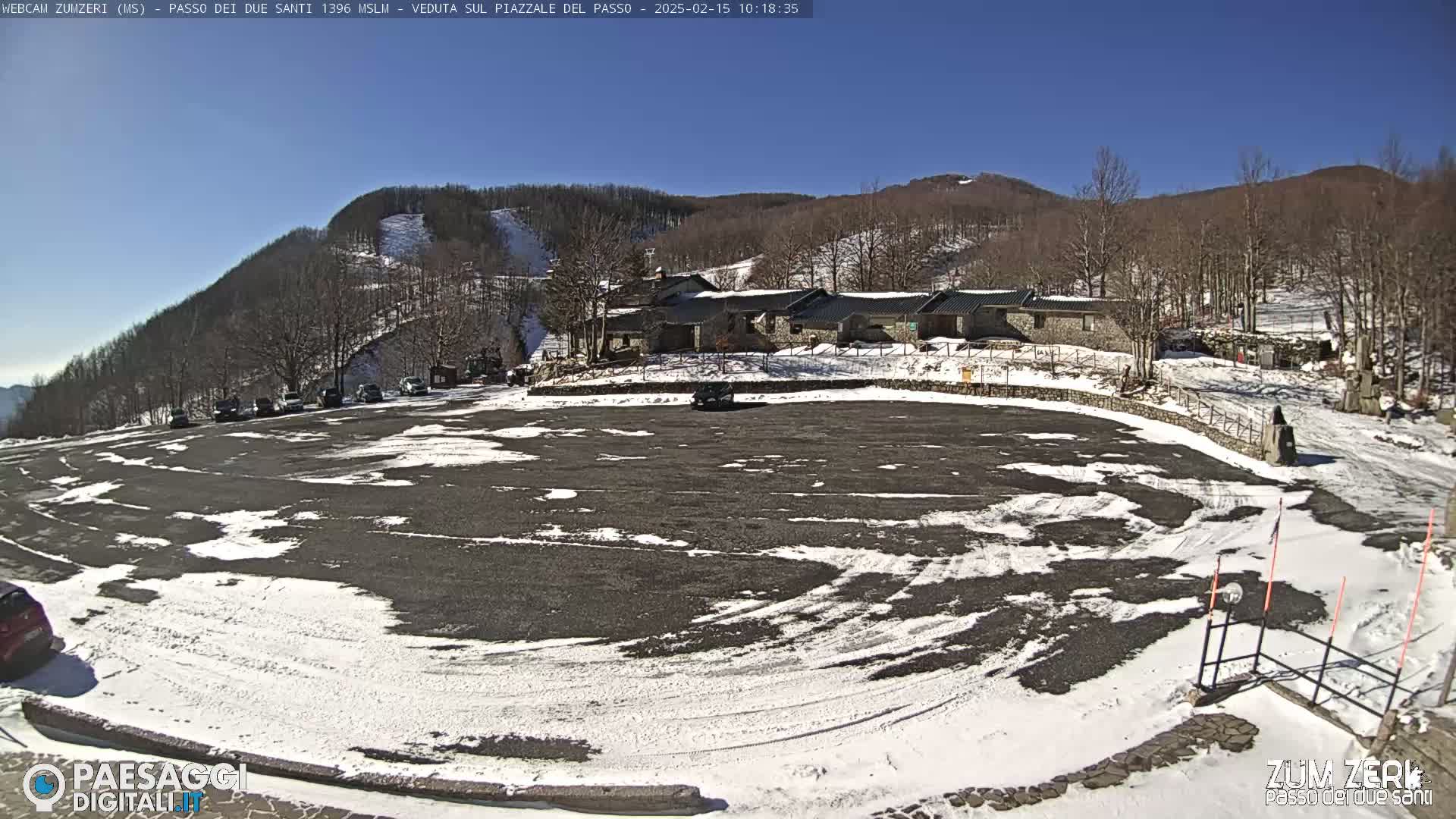 A mostly empty asphalt parking lot with patches of snow is situated in front of a stone building complex nestled against a snow-dusted mountainside under a clear blue sky.