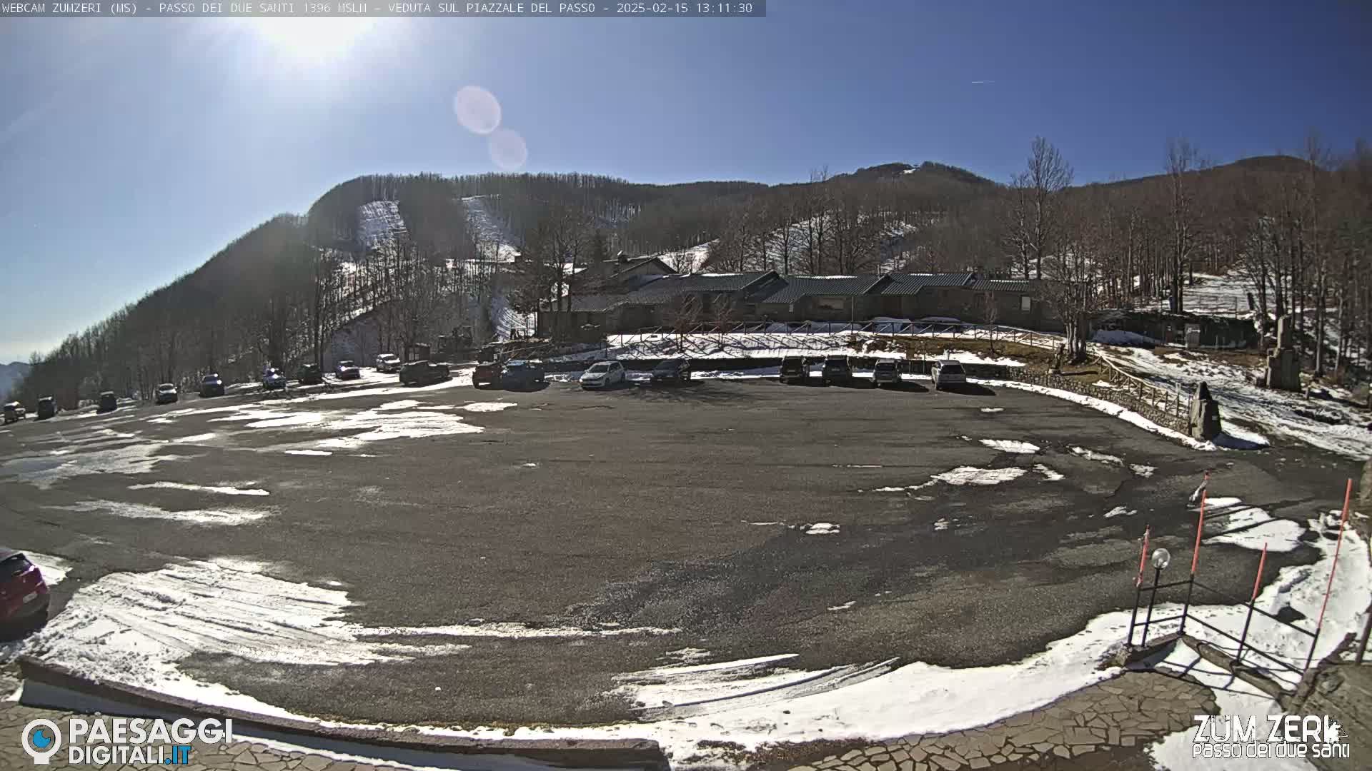 A mostly empty parking lot with some cars, patches of snow, and a building nestled in a snow-dusted mountainside under a clear blue sky.