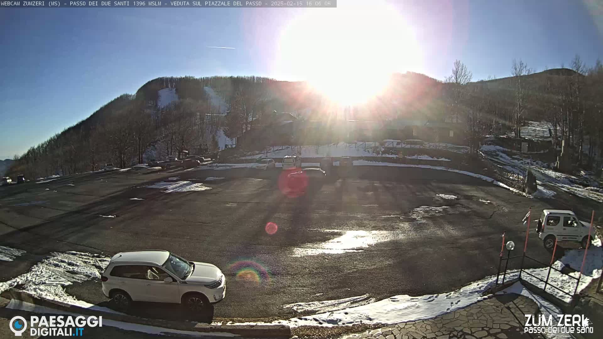 A sunny, partially snow-covered parking lot with several cars and a building in the background, situated on a mountainside.
