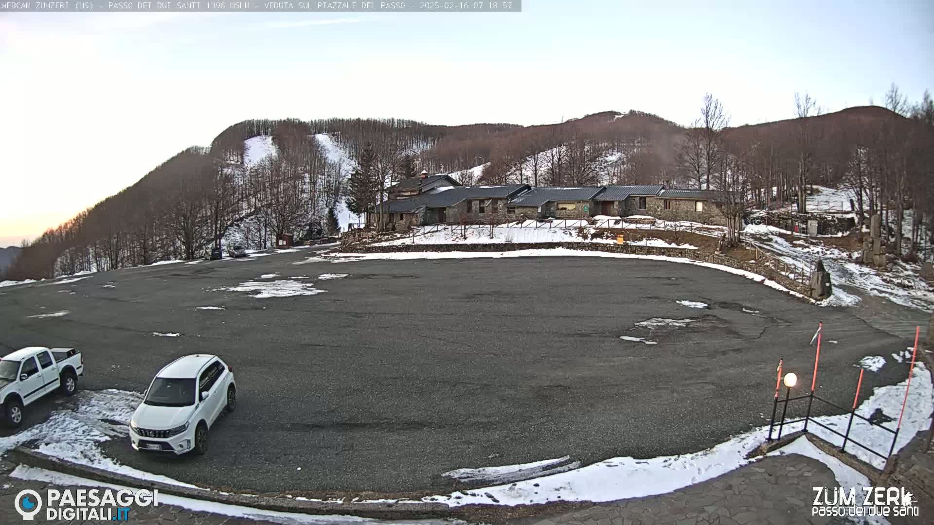 A large, mostly empty asphalt parking lot sits below a stone building complex nestled among snow-dusted, leafless trees on a lightly snow-covered hillside under a clear, bright sky.