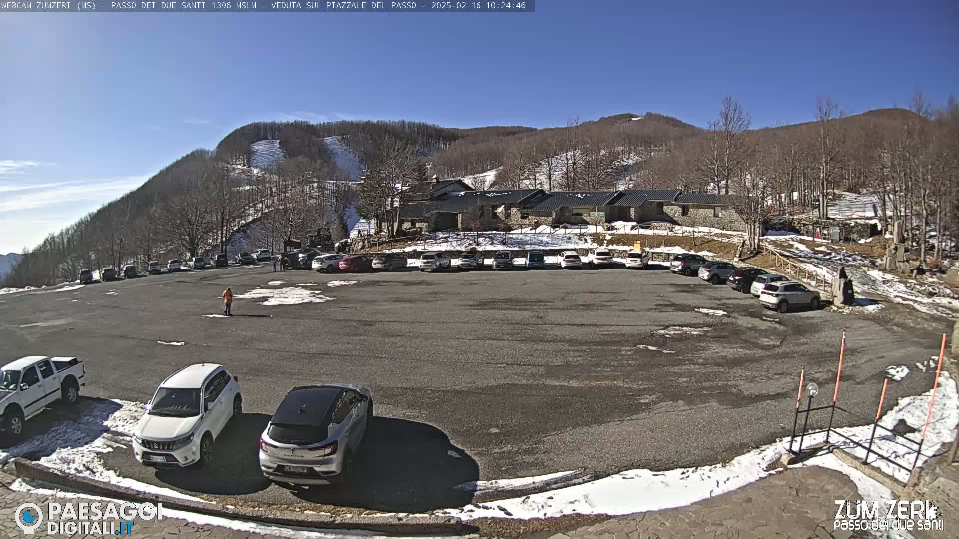 A paved parking lot filled with cars sits in front of a stone building, with a snow-covered mountain and clear sunny skies in the background.