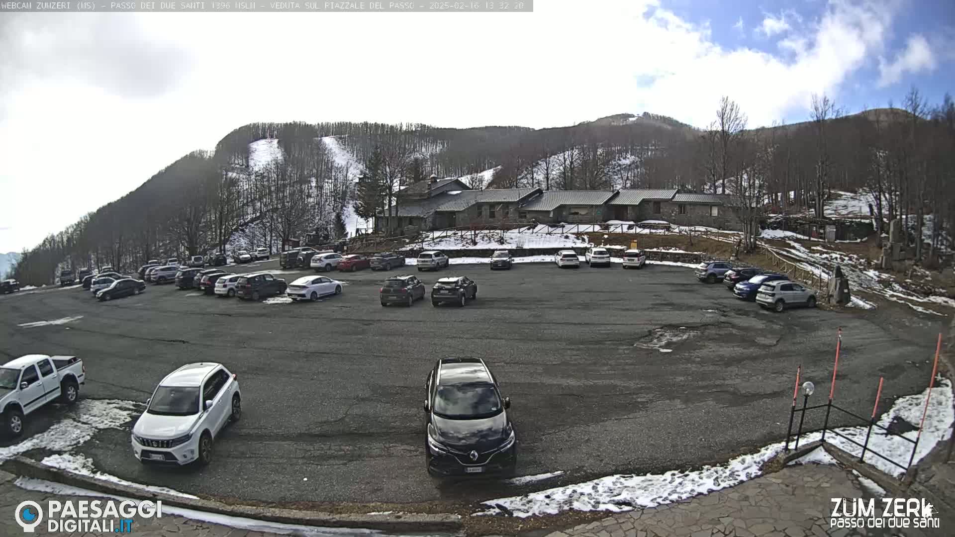 A parking lot filled with cars sits below a building in a mountain area with some snow on the ground and hillsides under a partly cloudy sky.